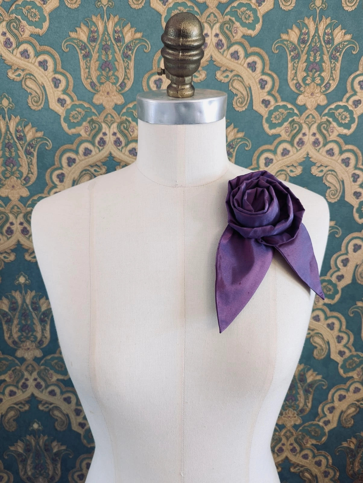 Broochella Petalo handmade silk flower brooch with petal detail in purple. Pinned to a mannequin. The background is green and gold damask wallpaper.