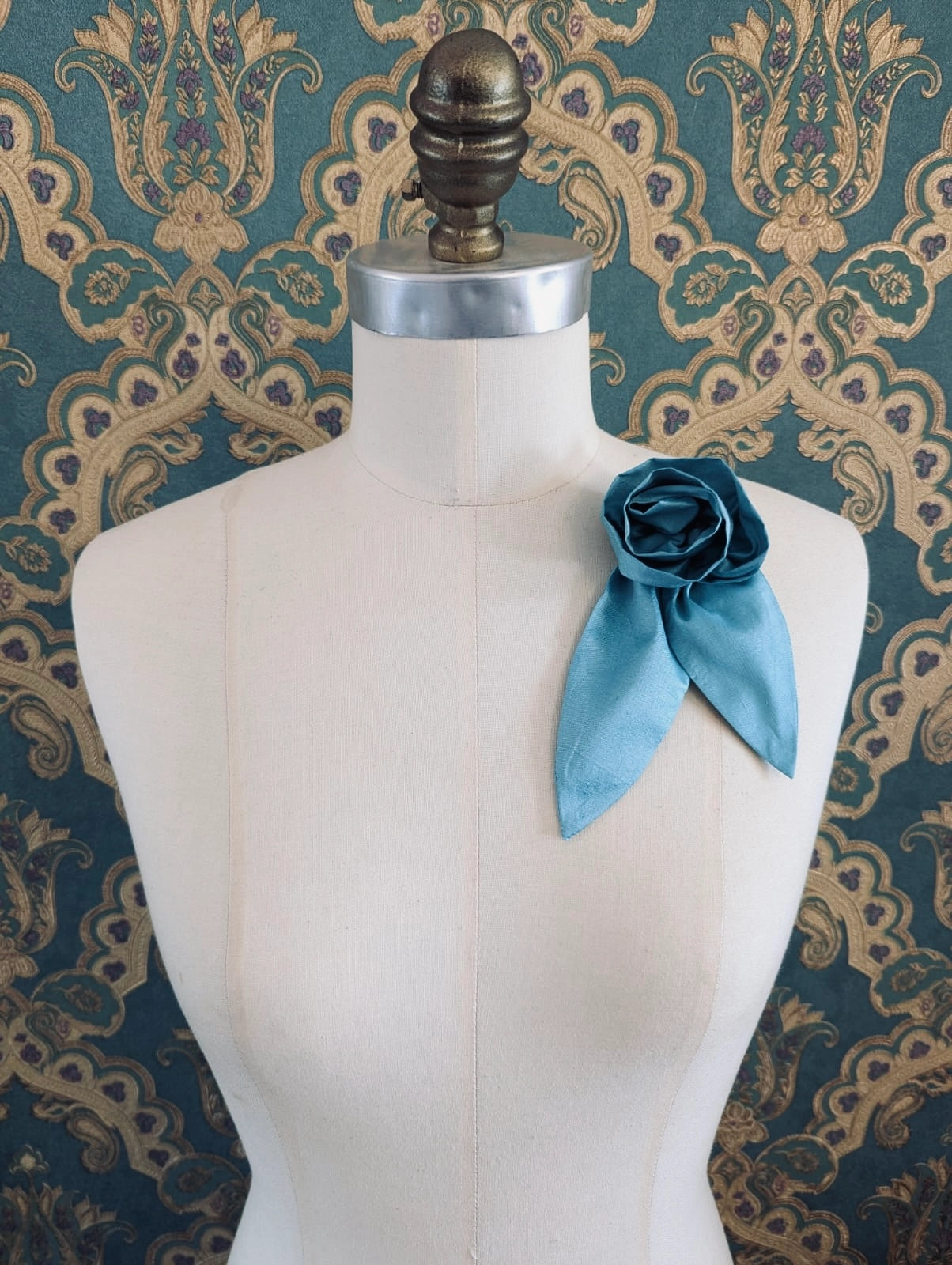 A Broochella Petalo blue, small, handmade silk  rose-shaped flower brooch with petals. It is displayed on a cream-coloured fabric dress form against a dark teal and gold damask patterned wallpaper background.