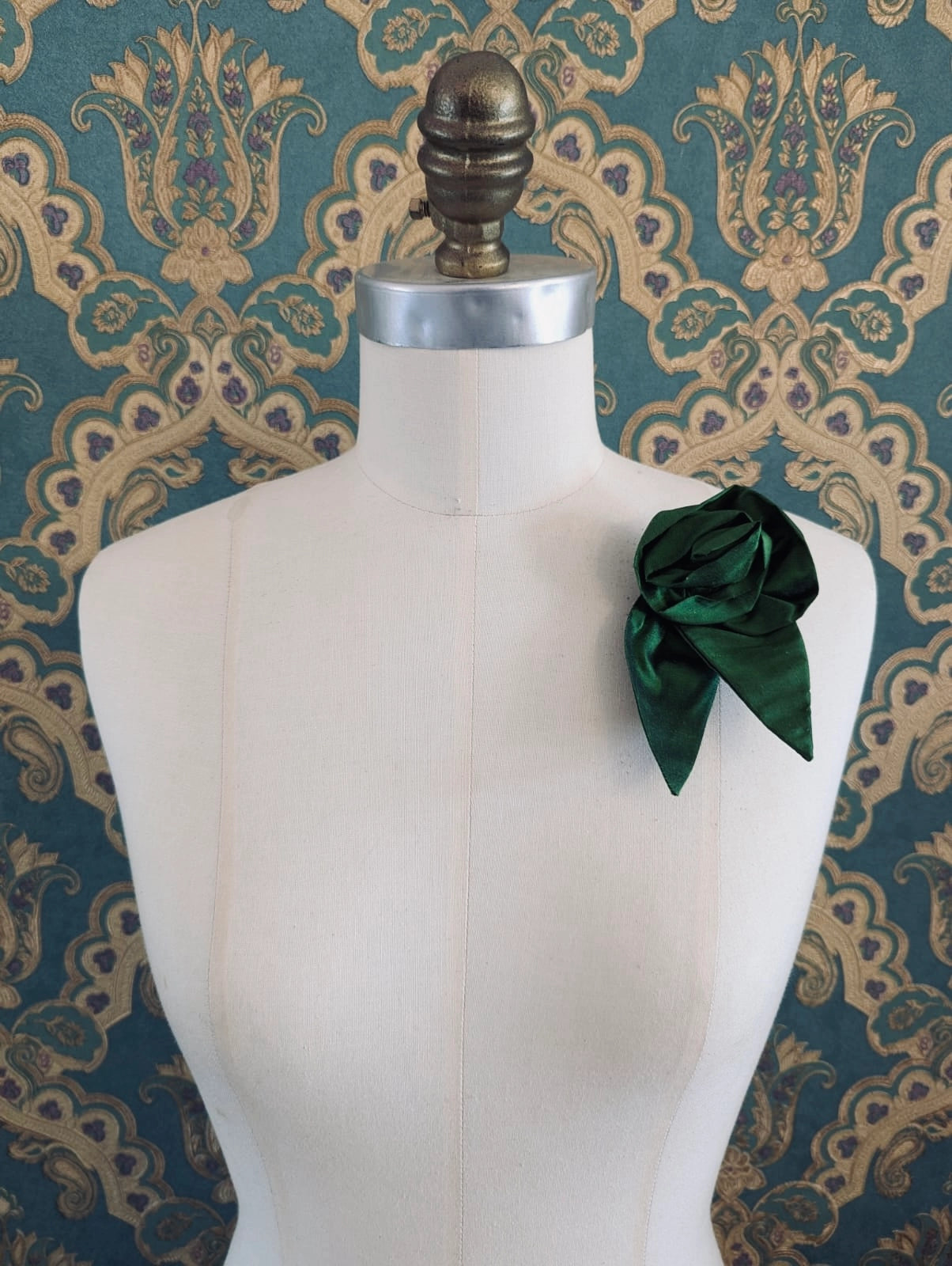 A Broochella Petalo green, small, handmade silk  rose-shaped flower brooch with petals. It is displayed on a cream-coloured fabric dress form against a dark teal and gold damask patterned wallpaper background.