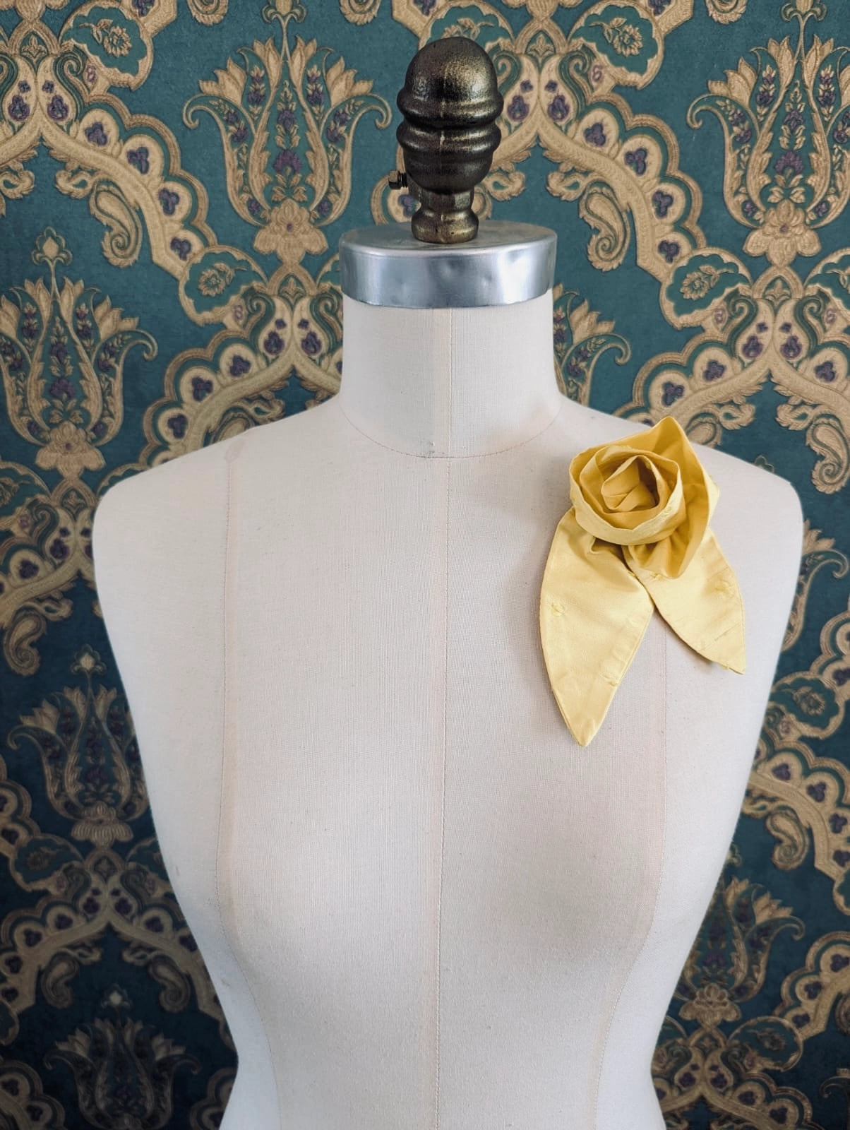 A Broochella Petalo yellow, small, handmade silk  rose-shaped flower brooch with petals. It is displayed on a cream-coloured fabric dress form against a dark teal and gold damask patterned wallpaper background.
