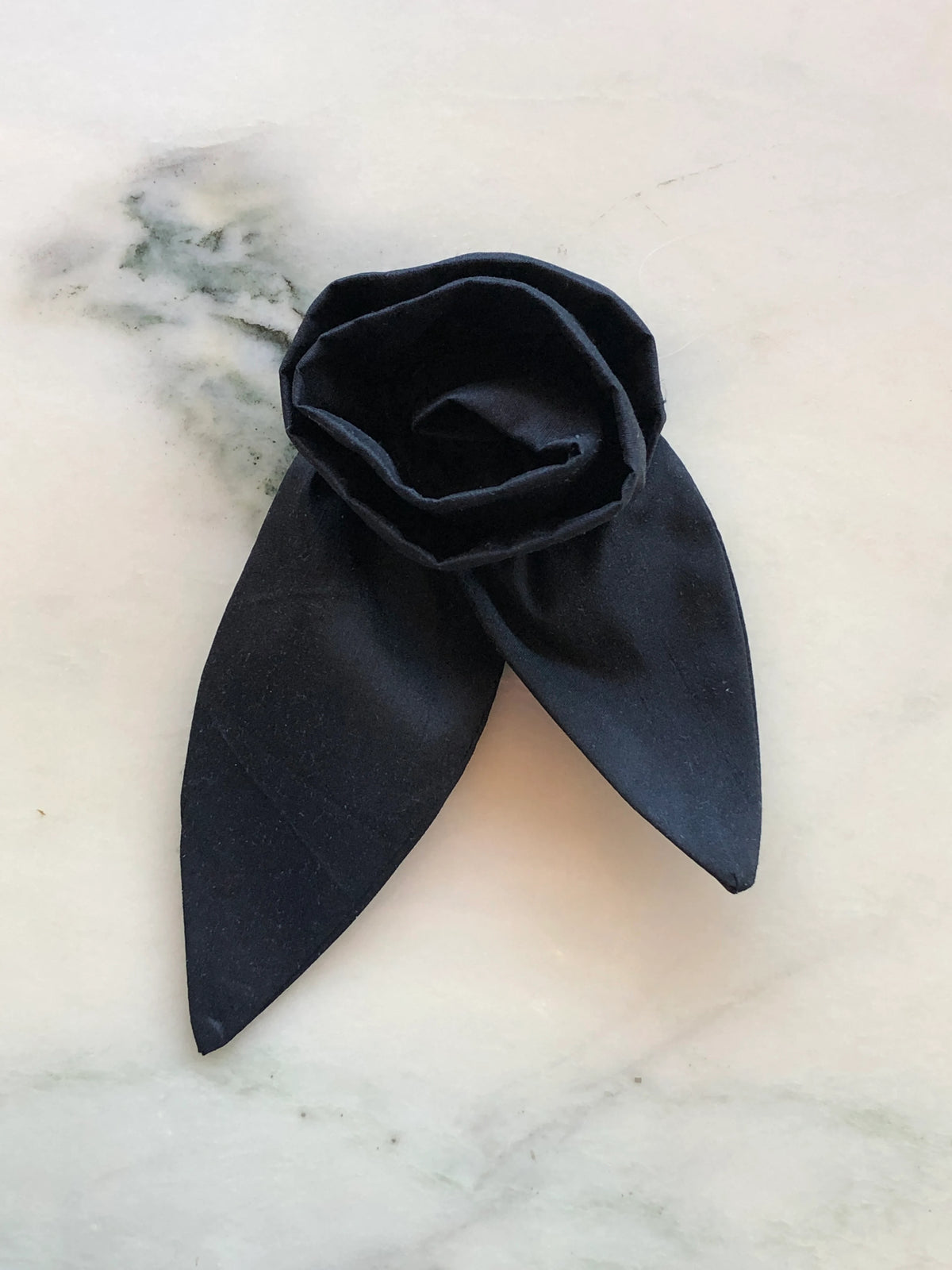 Broochella Petalo Small black silk flower brooch with sculptural rose petals and two pointed silk leaves, photographed on white marble with grey-green veining.