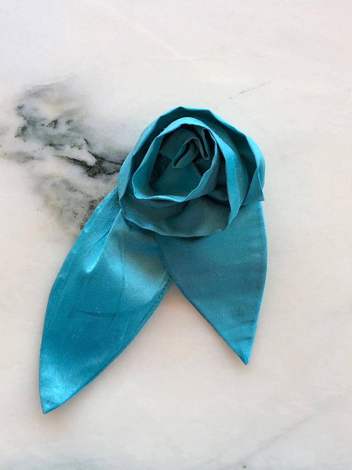 Broochella Petalo Small blue silk flower brooch with sculptural rose petals and two pointed silk leaves, photographed on white marble with grey-green veining.