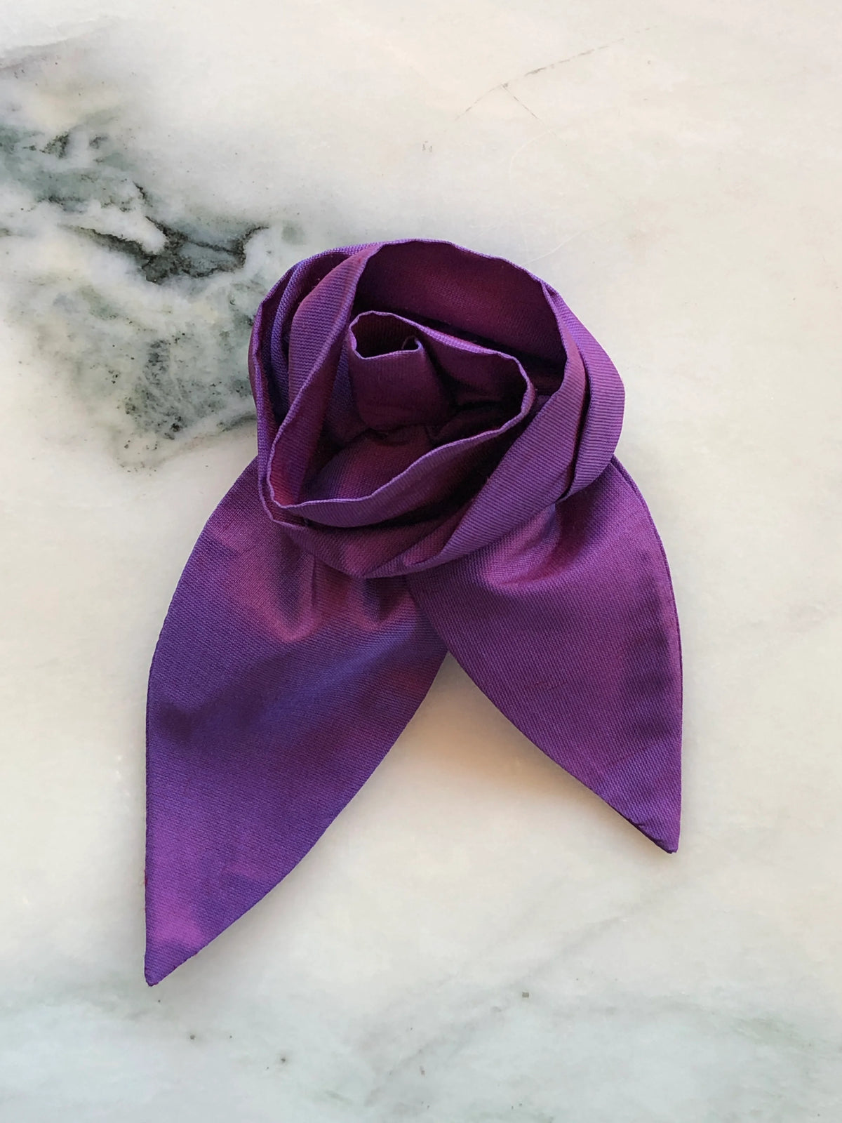 Broochella Petalo Small purple silk flower brooch with sculptural rose petals and two pointed silk leaves, photographed on white marble with grey-green veining.