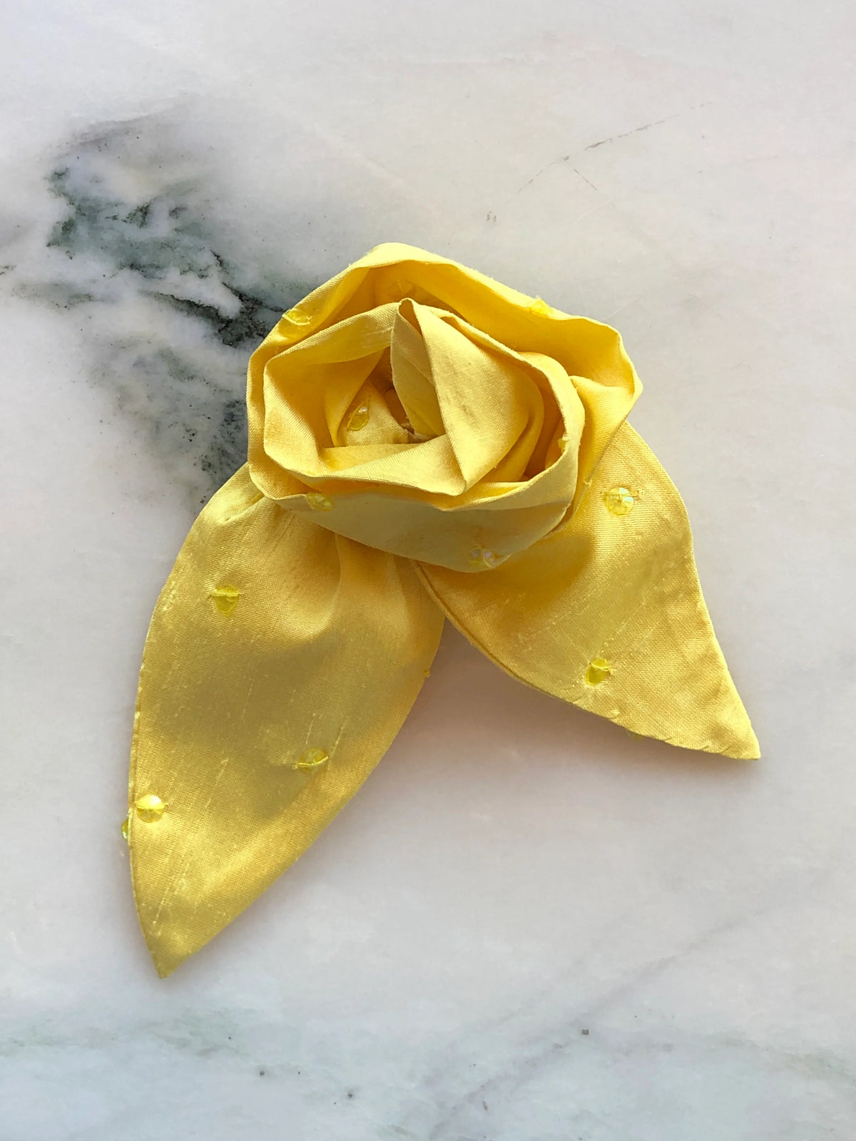 Broochella Petalo Men yellow silk flower brooch for men with sculptural rose petals, two pointed silk leaves and subtle embroidered details, photographed on white marble with grey-green veining.