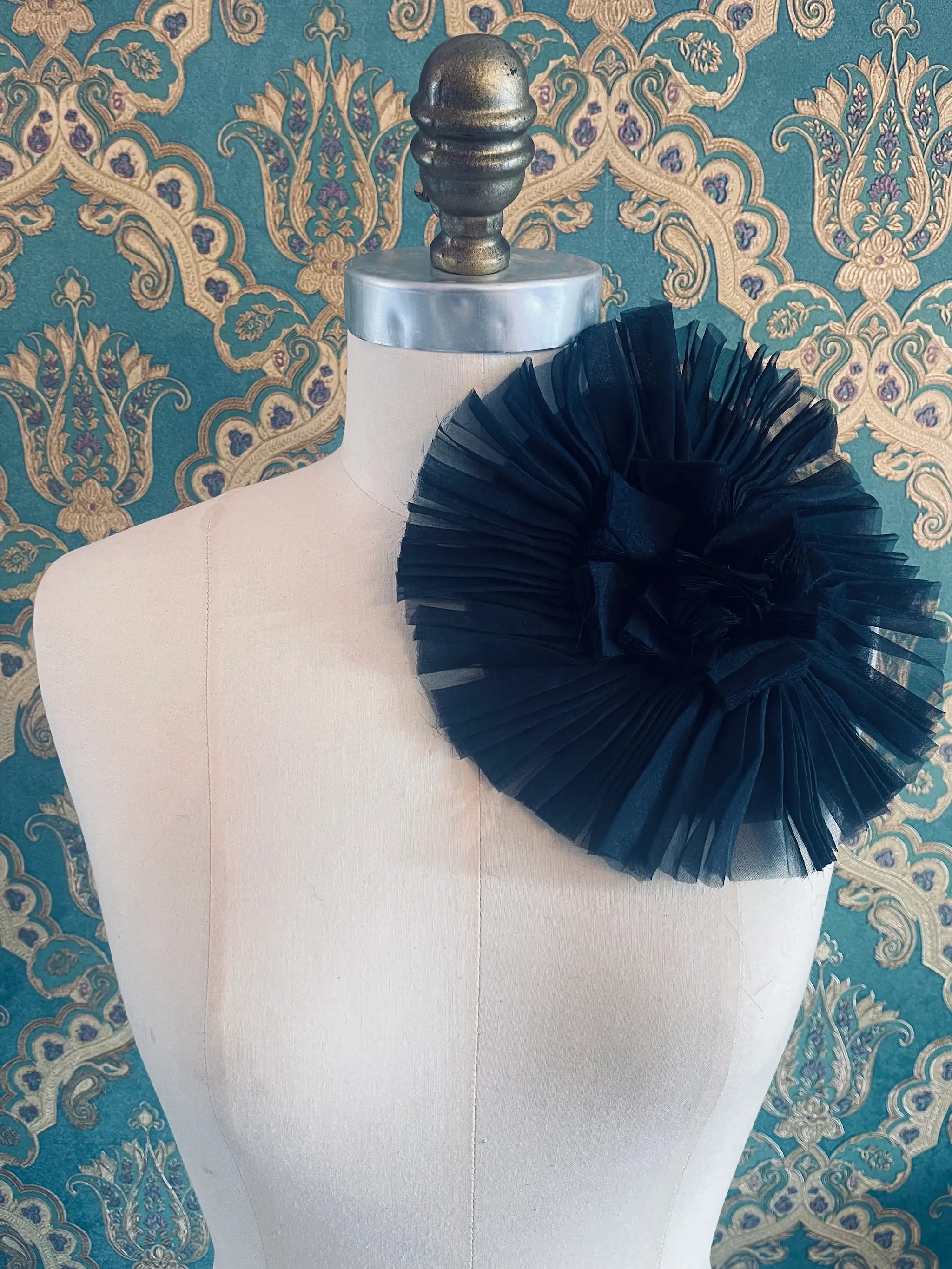 Broochella Pieghettato large handmade fabric flower brooch in black. Styled on a mannequin. The background is green and gold damask wallpaper