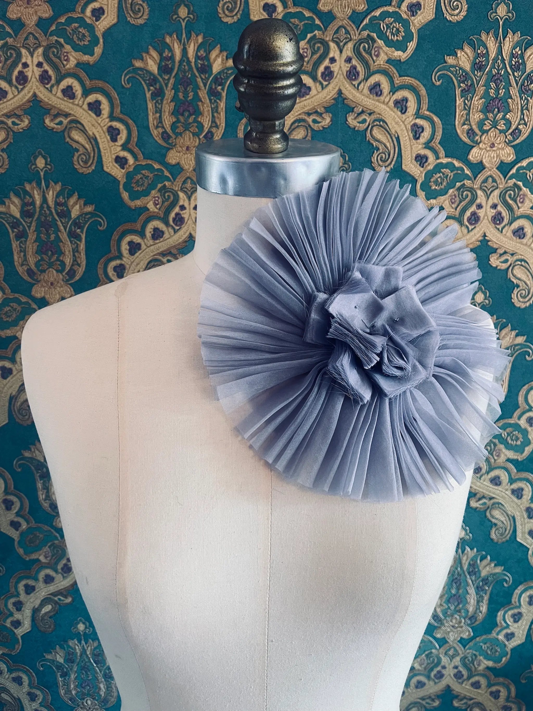 Broochella Pieghettato large handmade fabric flower brooch in lavender. Styled on a mannequin. The background is green and gold damask wallpaper