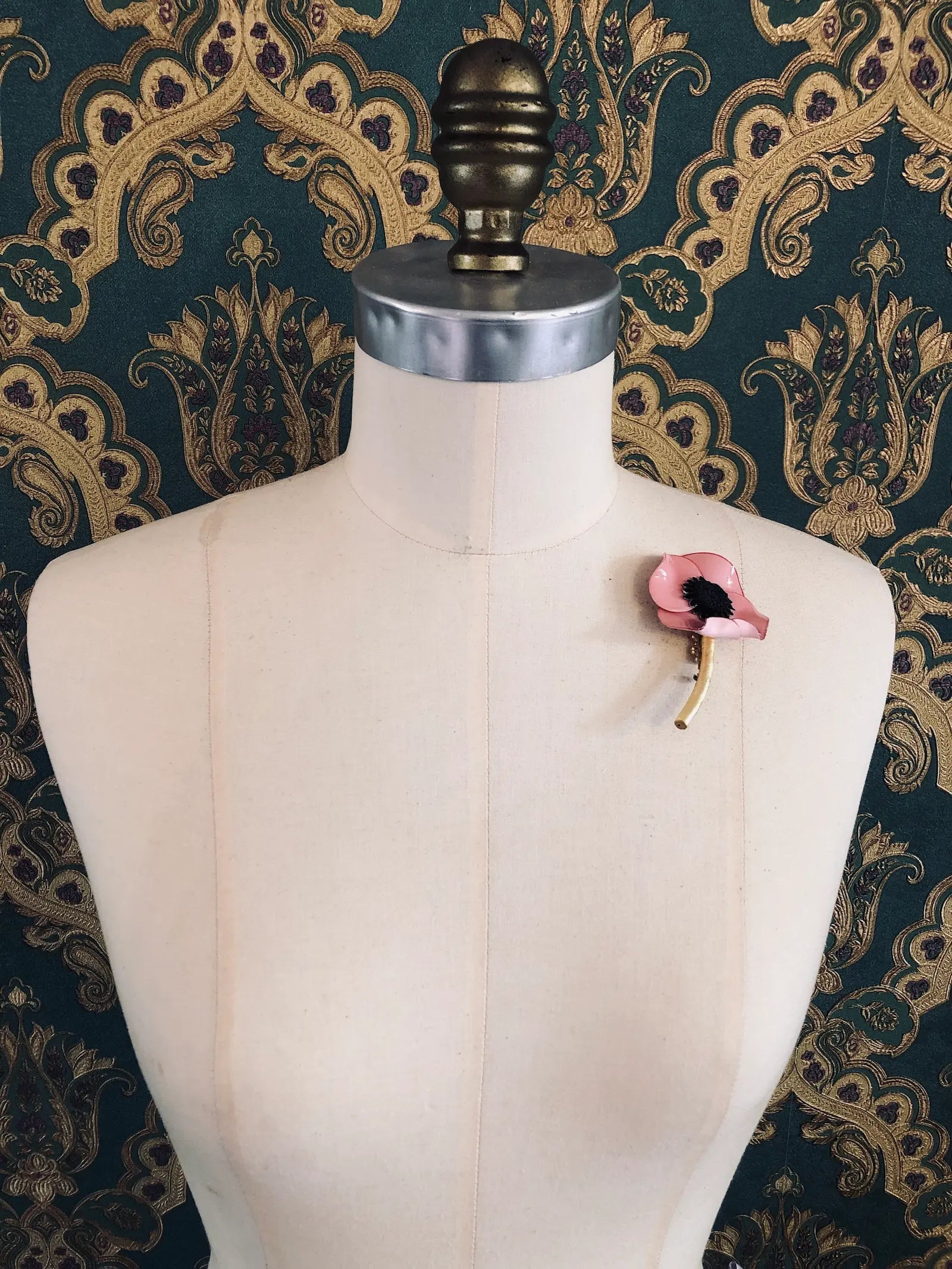 Broochella x Fabrice Paris pink moulded acetate poppy flower brooch in red. It is pinned to a mannequin. The background is green and gold damask wallpaper. 