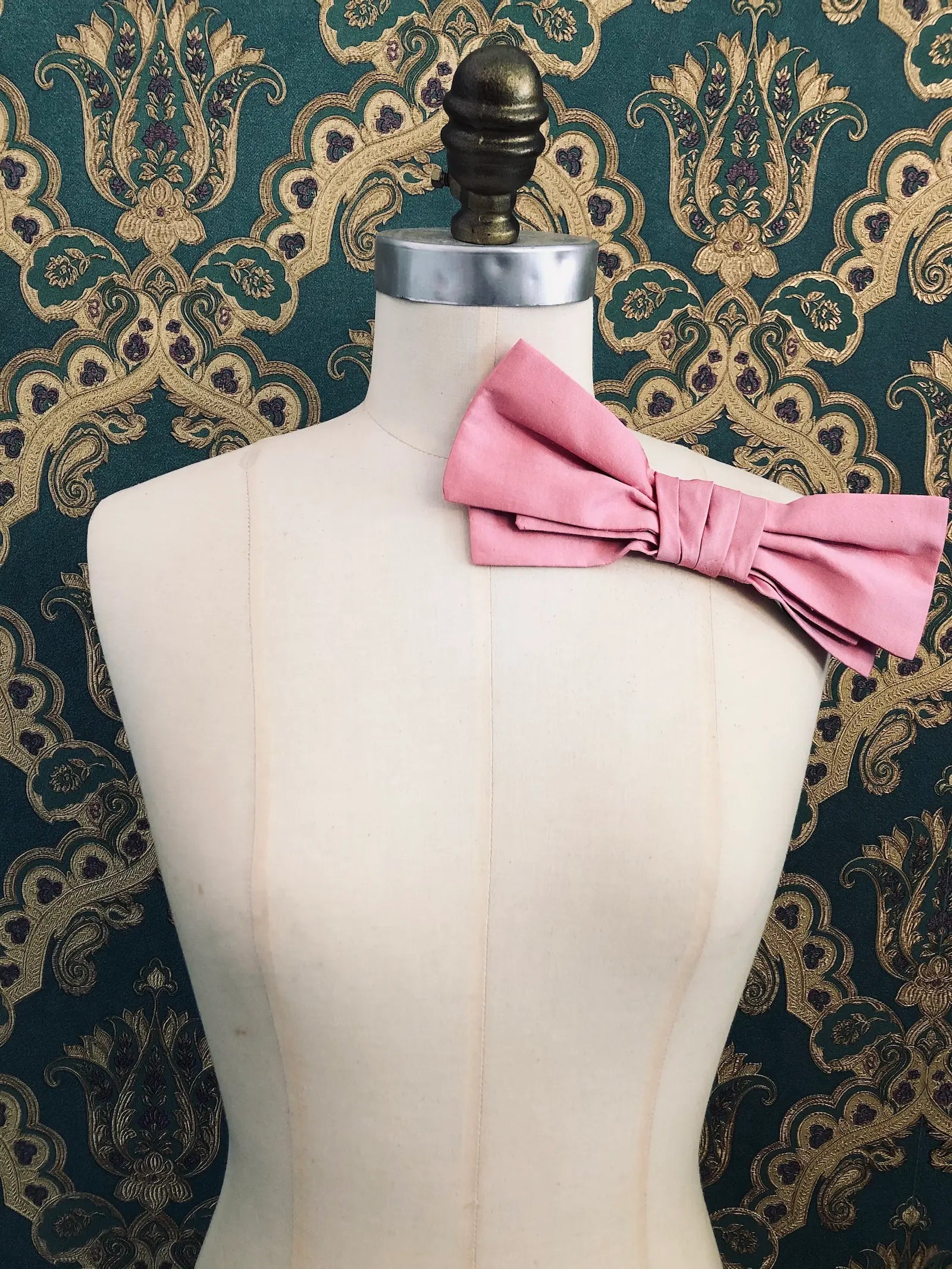 Broochella Marie Antoinette handmade, designer silk bow brooch in pink. It is pinned to a mannequin. The background is green and gold damask wallpaper.