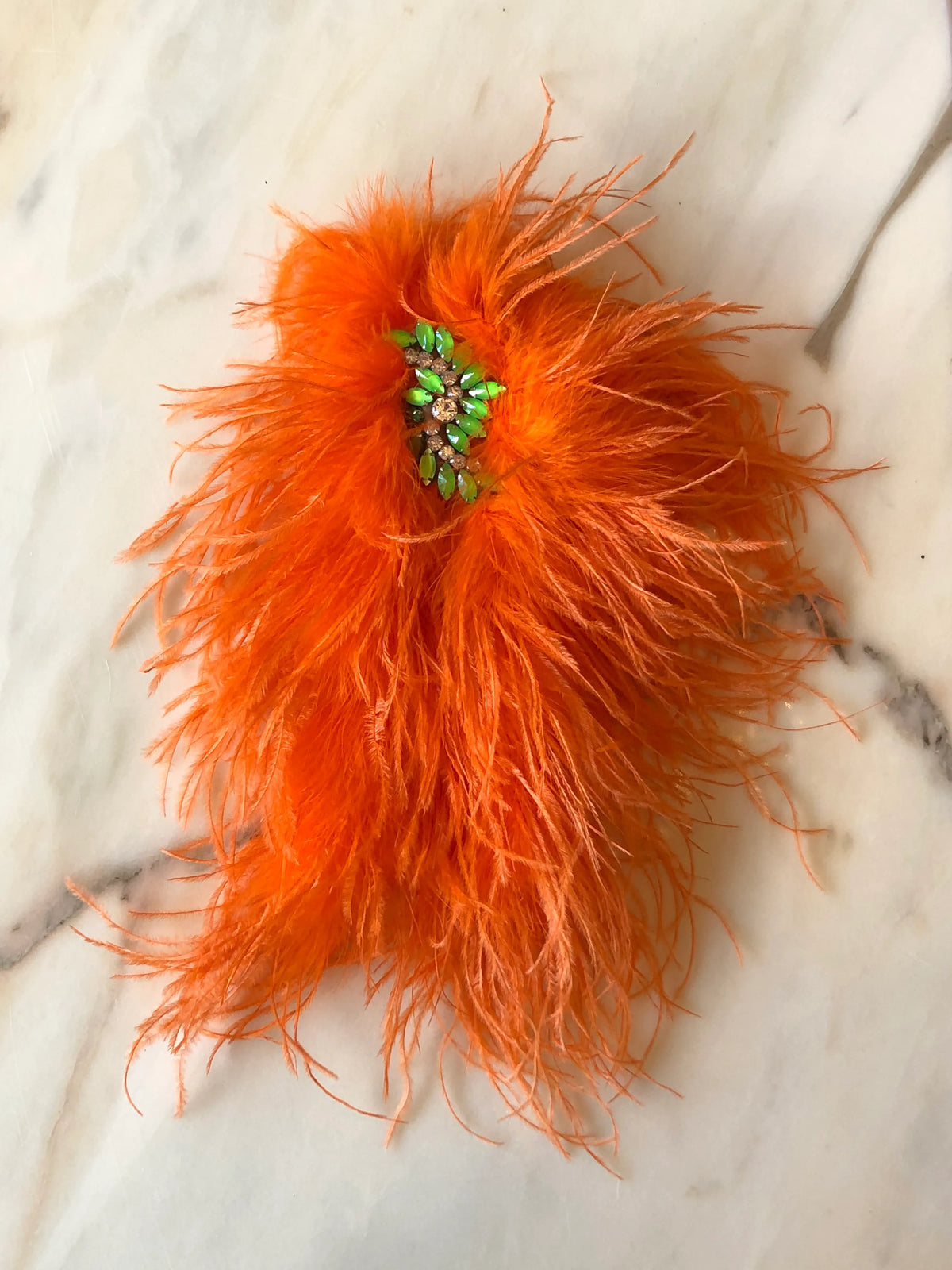 Broochella Piuma Neon Feather Brooch Pin in vibrant neon orange ostrich feathers with a contrasting green jewelled centre, photographed on a white marble background.