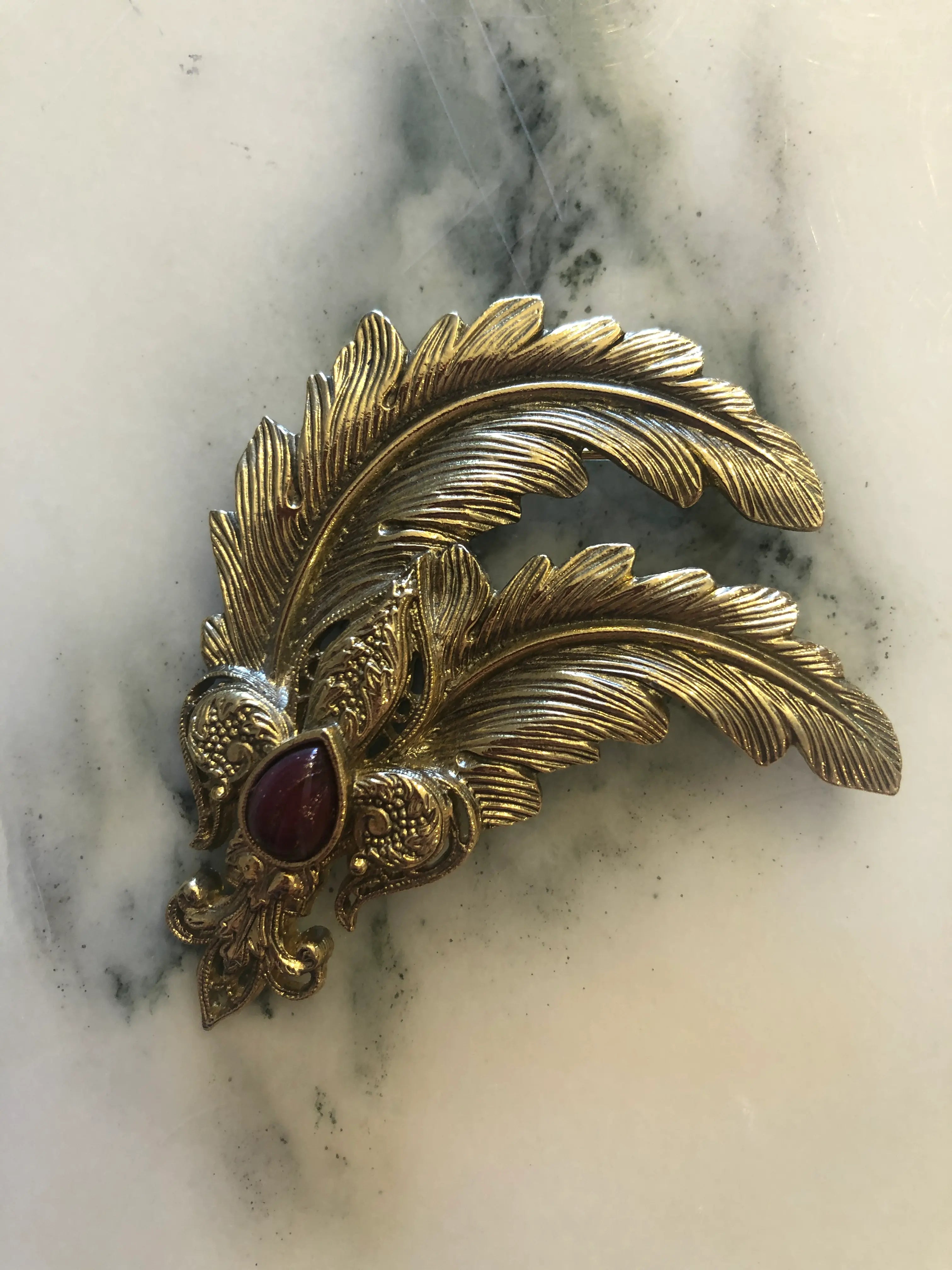 A Broochella plum imperiale ornate vintage brooch featuring a gold-toned feather design with a central teardrop-shaped dark red gemstone, displayed on a white and gray marbled surface.