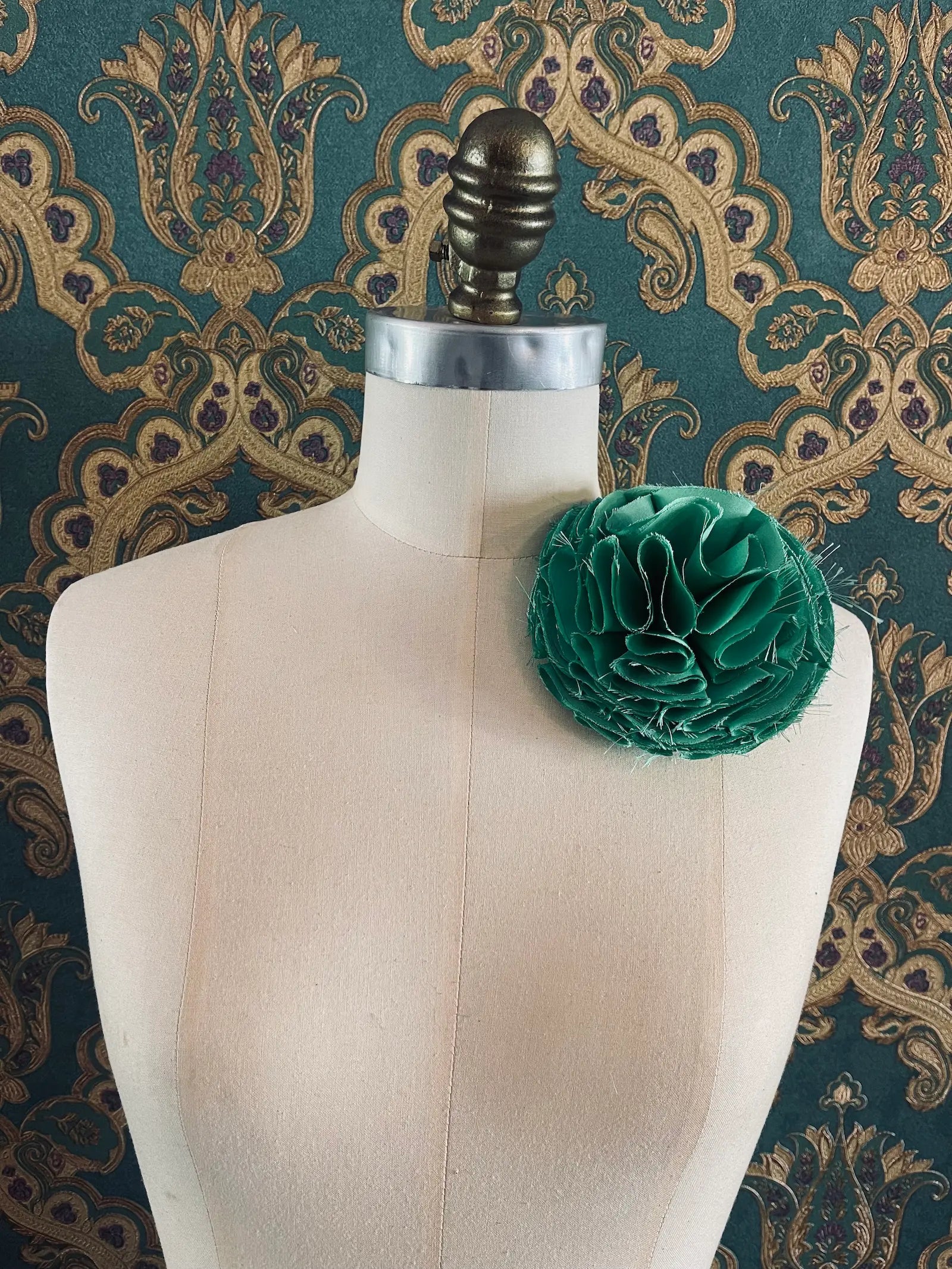 Broochella Pom Pon handmade designer flower brooch in green. Pictured on a mannequin. The background is green and gold damask wallpaper.