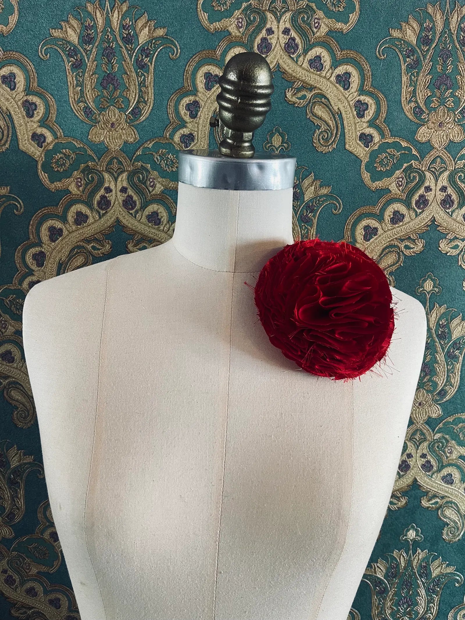 Broochella Pom Pon handmade designer flower brooch in red. Pictured on a mannequin. The background is green and gold damask wallpaper.