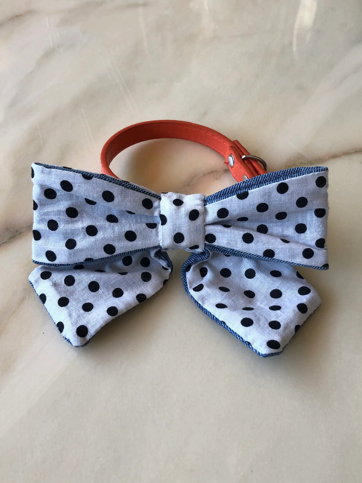Broochella Poochella white cotton dog bow with black polka dots, blue denim edging and pointed tails, displayed with a coral orange pet collar on a cream marble background with soft beige and grey veining.