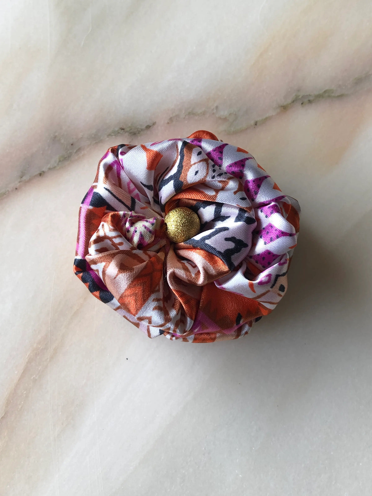 Broochella Poochella Bougainville Capri satin pet flower featuring layered, gathered petals in a vibrant pink, orange, cream and black abstract print with a textured gold centre, photographed from above on a cream marble background with soft grey and blush veining.