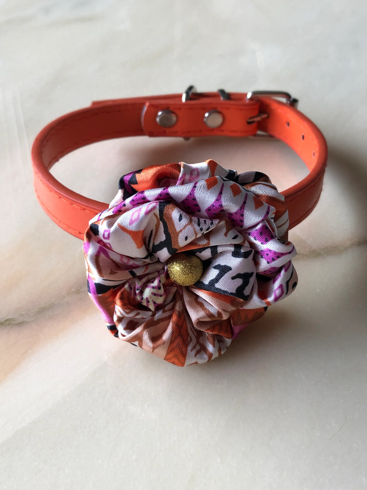 Broochella Poochella Bougainville Capri pet flower collar featuring a gathered satin flower with pink, orange, cream and black abstract print, a textured gold centre and an adjustable orange collar with silver hardware, photographed on a polished cream marble background with subtle grey veining.