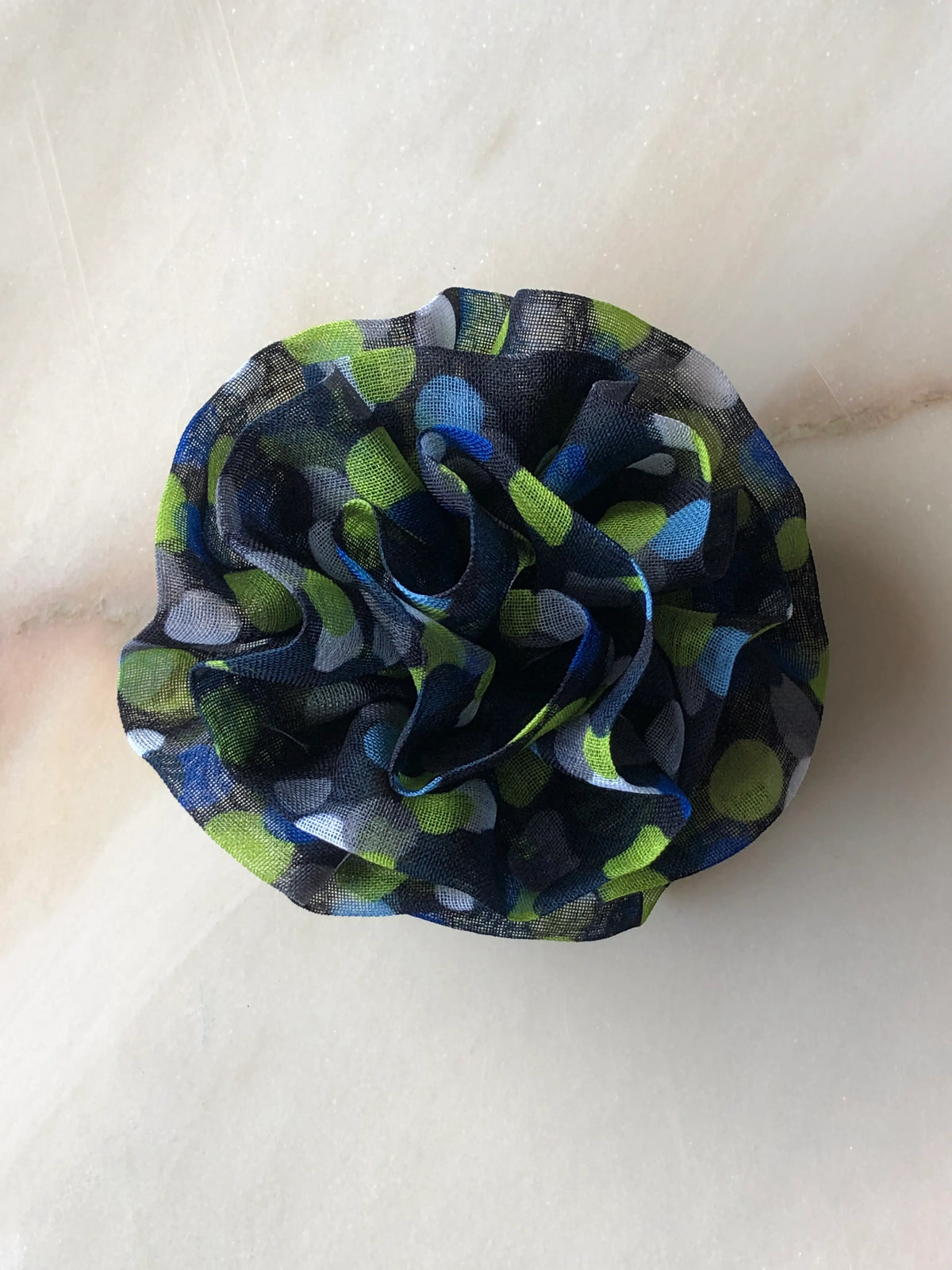 Broochella Poochella Brassica blue pet flower featuring a sculptural rosette of gathered sheer navy mesh with lime-green, cobalt-blue, pale-blue and grey polka dots, photographed from above on cream marble with subtle grey veining.