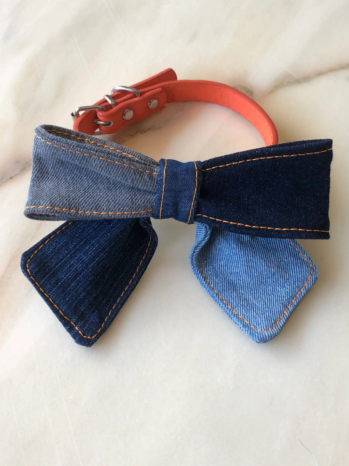 Broochella Poochella denim dog bow featuring contrasting light blue, charcoal and dark indigo denim panels, orange stitching and pointed tails, attached to an orange pet collar with silver hardware, photographed on polished cream marble with subtle beige and grey veining.