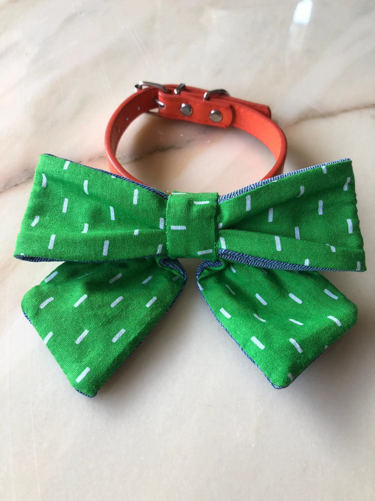 Broochella Poochella vibrant green cotton dog bow featuring a playful white dash print, gathered centre, contrasting blue denim edging and two pointed tails, attached to an orange pet collar with silver hardware, photographed on polished cream marble with subtle beige and grey veining.