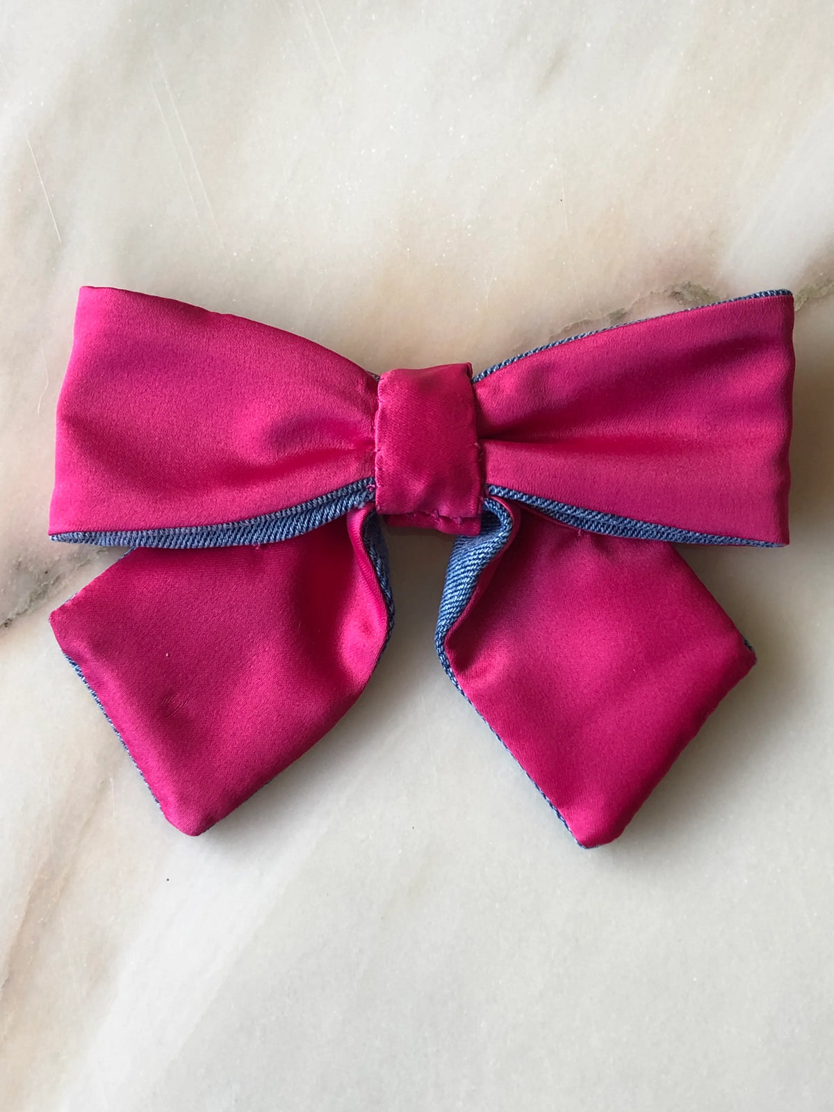 Broochella Poochella pink satin pet bow featuring lustrous fuchsia pink fabric, contrasting blue denim trim, structured loops, a gathered centre and two pointed tails, photographed on polished cream marble with soft beige and grey veining.