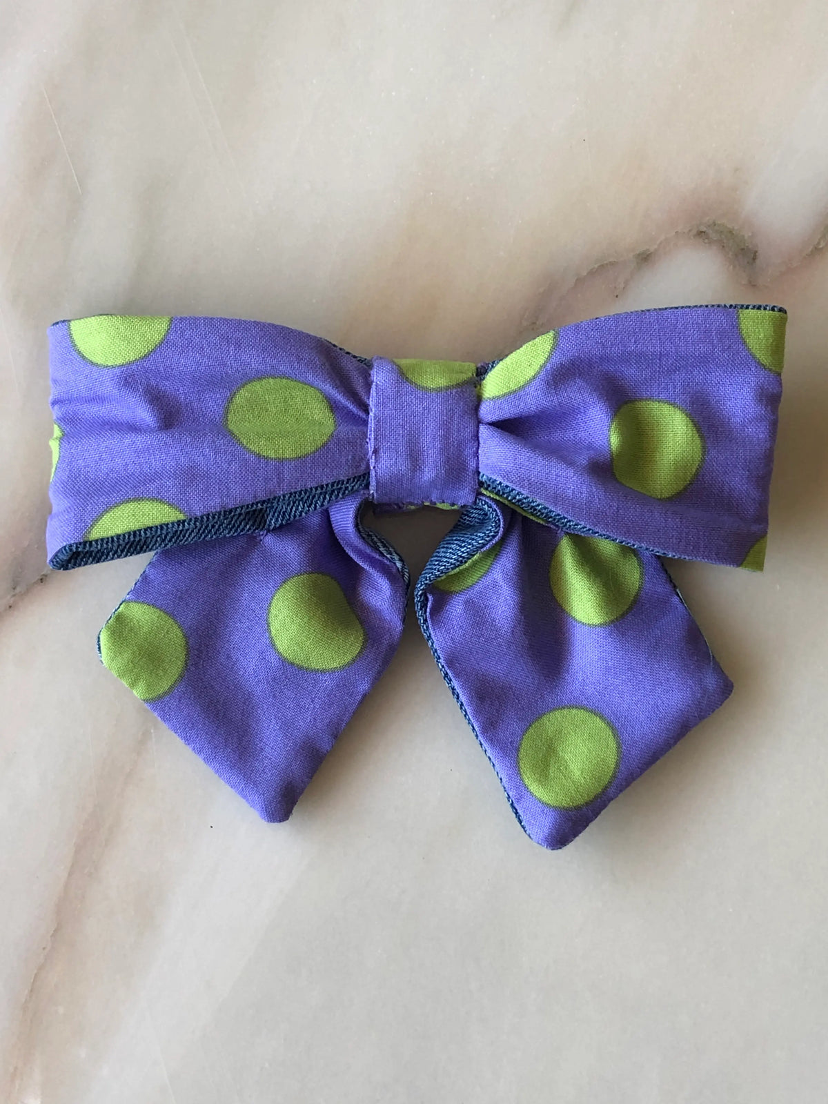 Broochella Poochella purple cotton pet bow featuring oversized lime green polka dots, structured loops, a gathered centre, pointed tails and blue denim edging, displayed on a polished cream marble background with soft beige and grey veining.