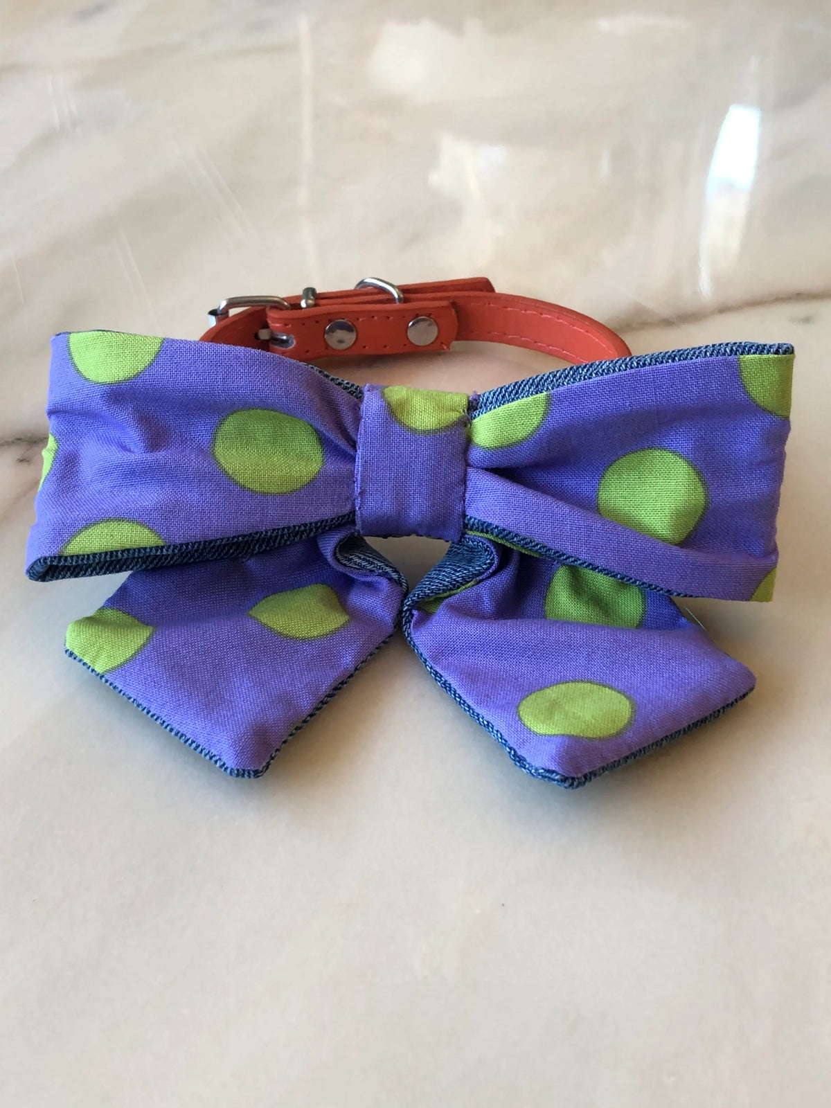 Broochella Poochella purple cotton dog bow with large lime green polka dots, contrasting blue denim edging and pointed tails, attached to an orange pet collar with silver hardware, photographed on a polished cream marble background with subtle beige and grey veining.