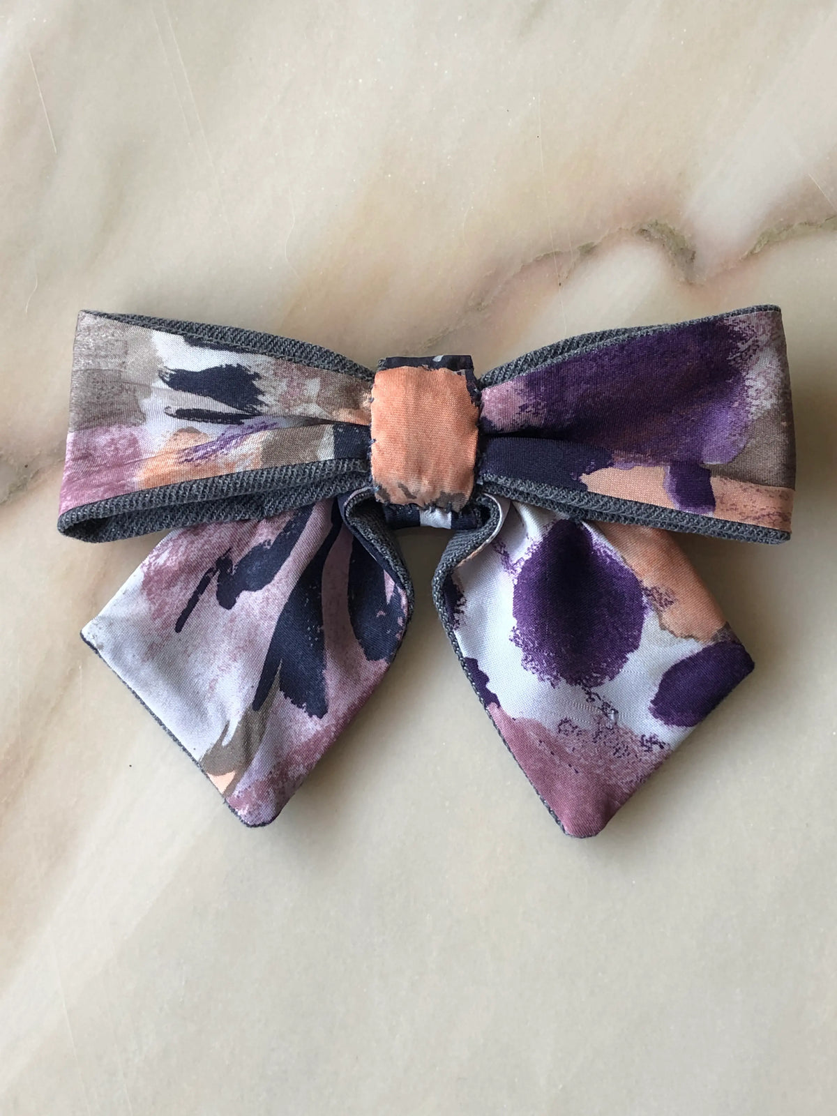 Broochella Poochella purple print pet bow featuring an artistic abstract brushstroke pattern in deep purple, mauve, blush pink, peach and charcoal, finished with grey denim trim, a gathered peach centre and two pointed tails, photographed flat on polished cream marble with delicate beige and grey veining.