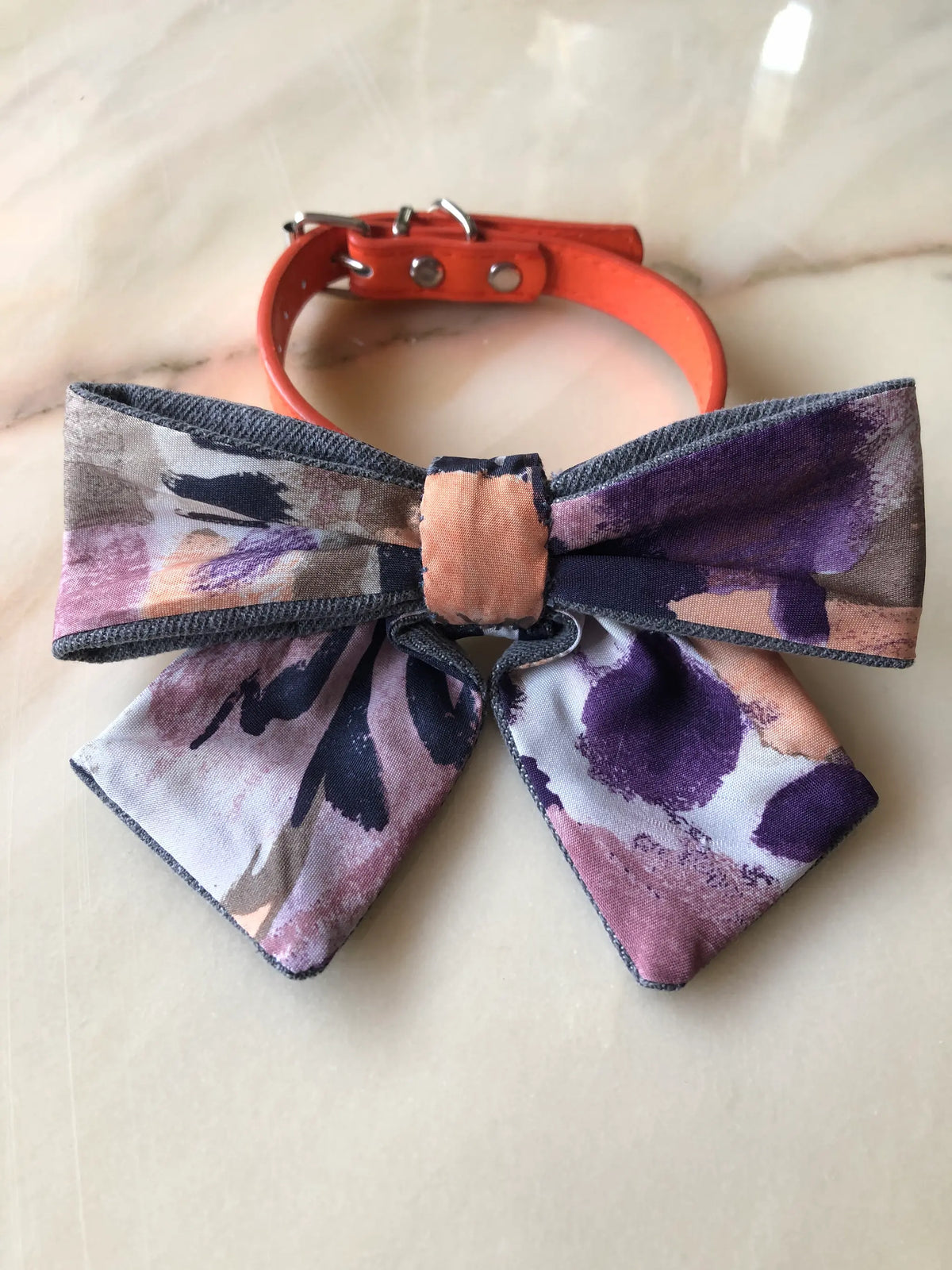 Broochella Poochella purple print dog bow featuring an abstract watercolour pattern in plum, lavender, blush pink, peach and charcoal, with contrasting grey denim edging and pointed tails, attached to an orange pet collar with silver hardware, photographed on polished cream marble with subtle beige and grey veining.