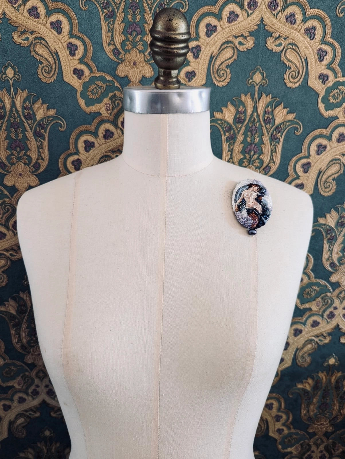 Broochella oval hand-beaded portrait of aphrodite nude figure in shades of blue and brown. Pinned on a mannequin. The background is green and gold damask wallapaper