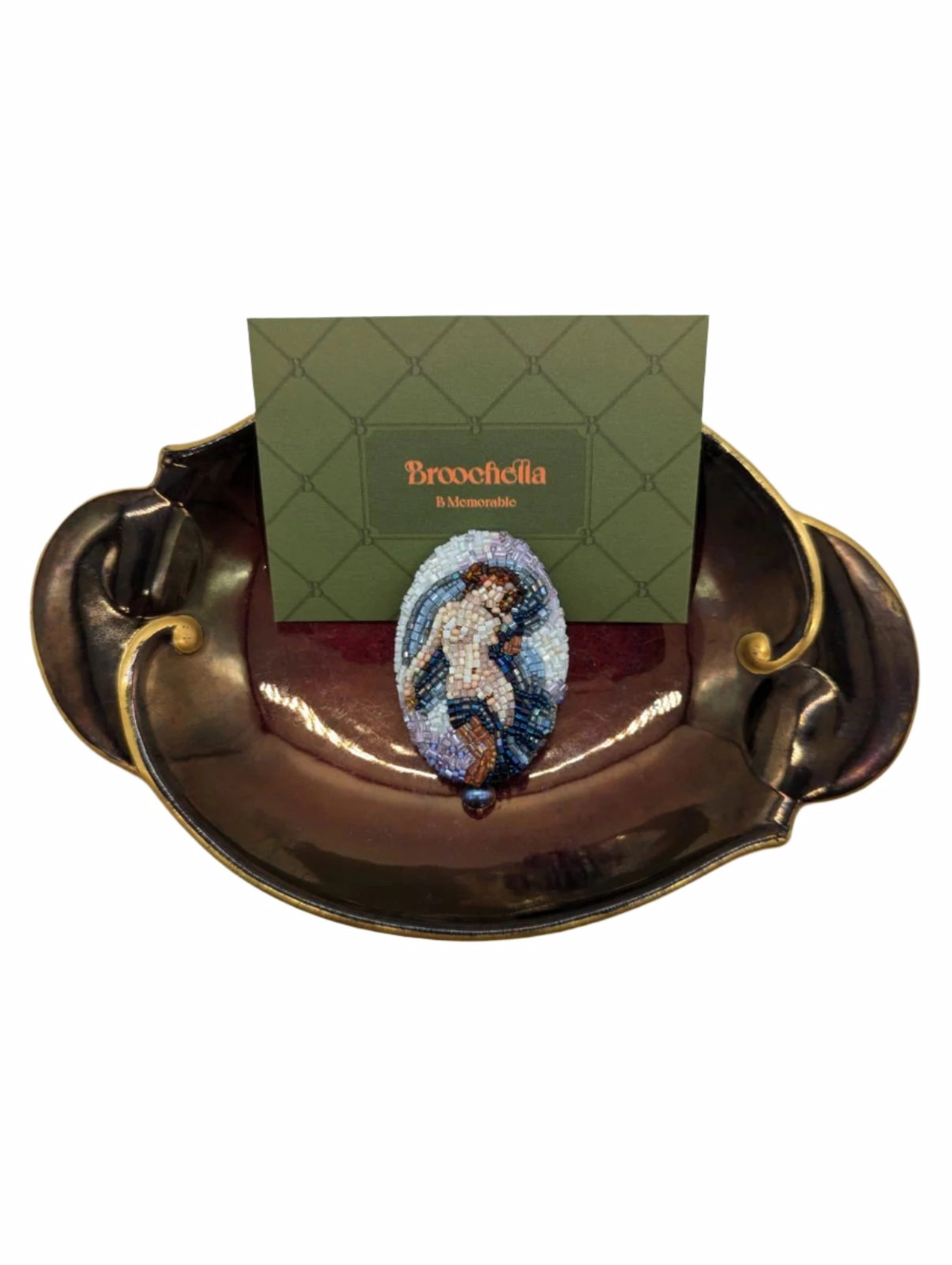 Broochella oval hand-beaded portrait of aphrodite nude figure in shades of blue and brown. it is placed on a burgundy and gold Wembley ware tray. There is a green broochella card behind the brooch. The background is white