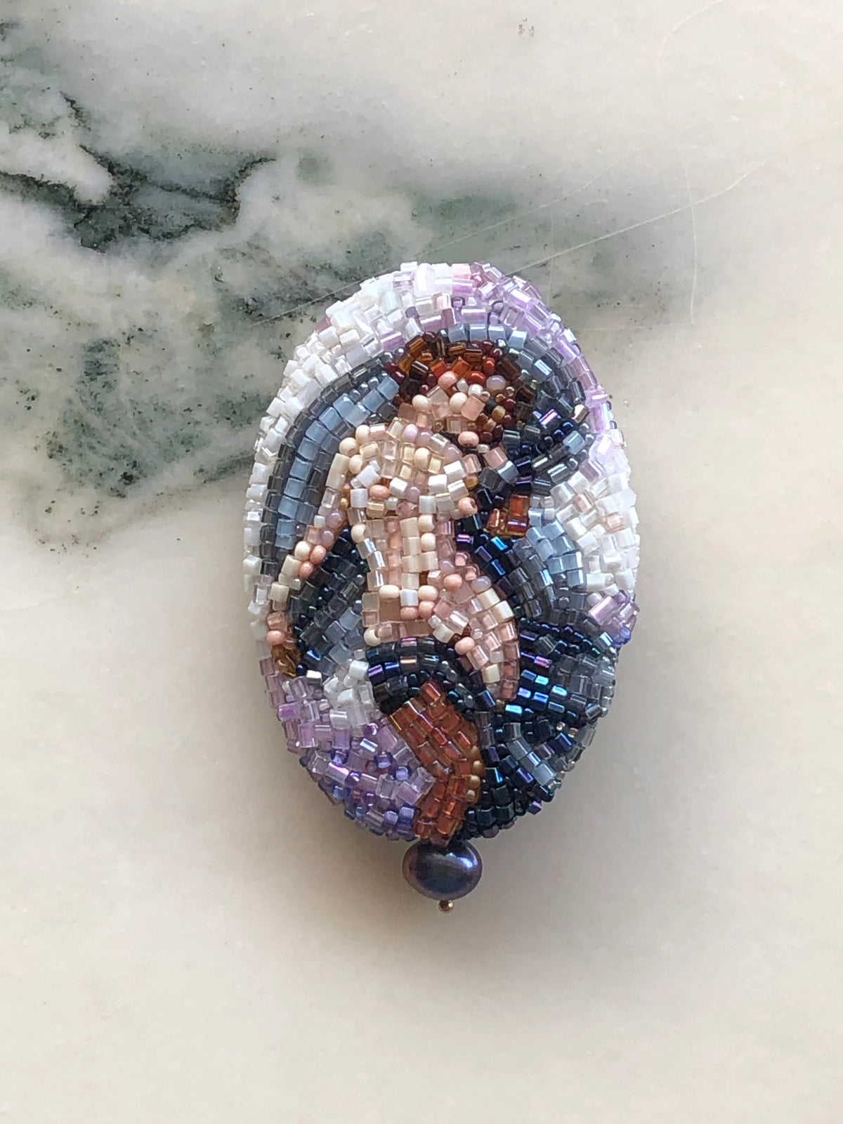 Broochella Portrait of Aphrodite Beaded Brooch featuring an intricately hand-beaded classical female figure in soft blush, ivory, silver, blue and bronze tones, photographed on a white marble background.