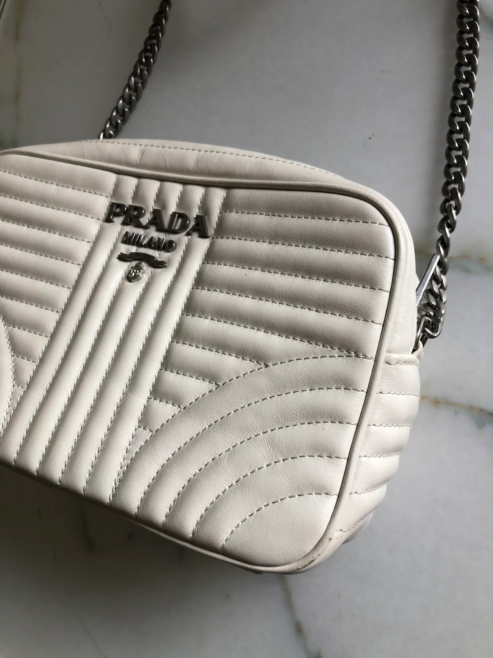 A close-up of a Broochella Prada Milano shoulder bag in off-white quilted leather with a chain strap, resting on a light marble surface.