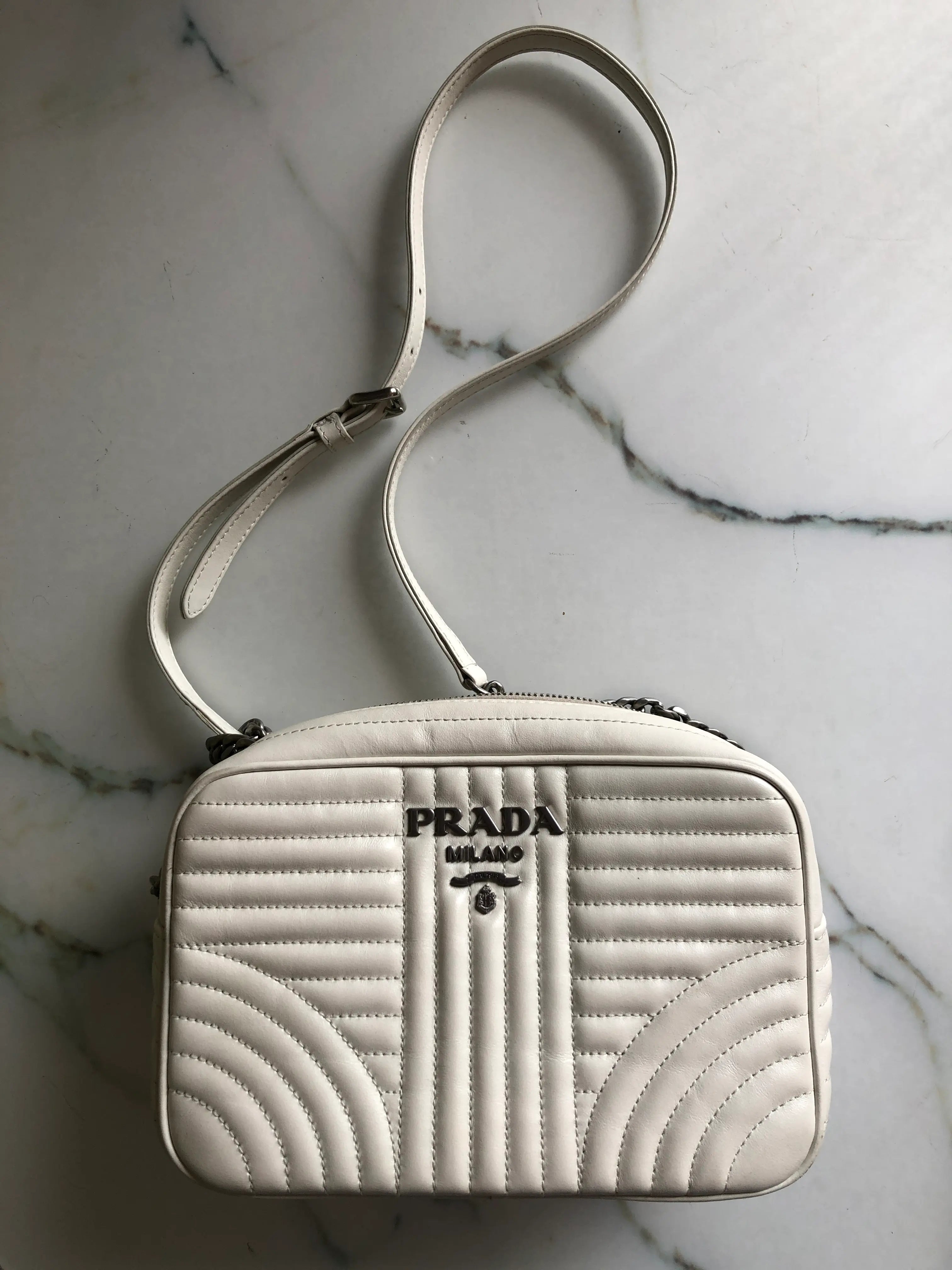 A white Broochella Prada Milano quilted crossbody bag with a matching strap, displayed on a white marble surface. The bag features a prominent Prada logo on the front.