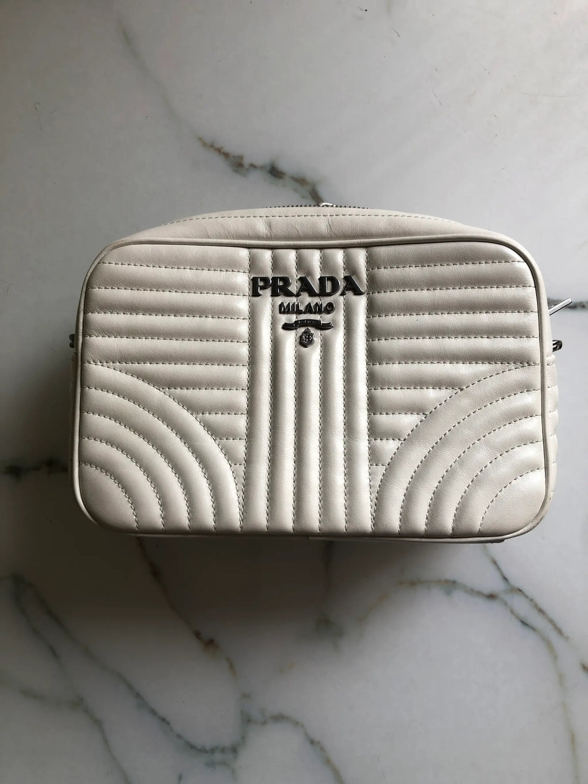 A Broochella Prada Milano bag in off-white quilted leather, featuring a central vertical pleated design, curved chevron patterns in the lower corners, and the black 'PRADA MILANO' logo on the front, resting on a white marble surface.