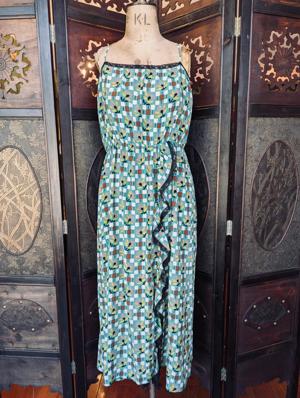 A Broochella authentic designer Prada sleeveless midi dress with a green, blue, and white checkered pattern and black ruffle trim, displayed on a vintage wooden mannequin torso in front of a dark wooden folding screen.