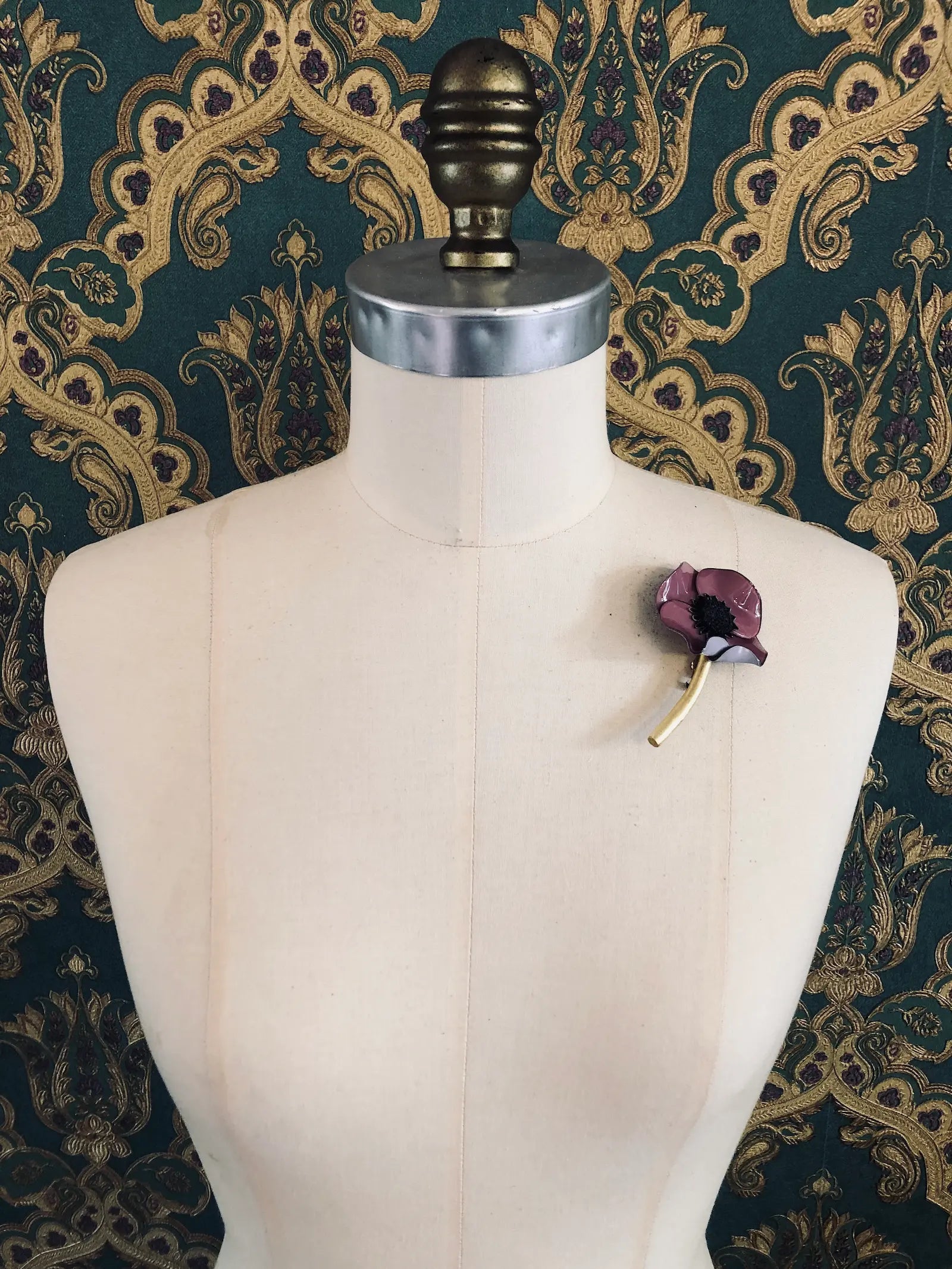 Broochella x Fabrice Paris purple moulded acetate poppy flower brooch in red. It is pinned to a mannequin. The background is green and gold damask wallpaper. 