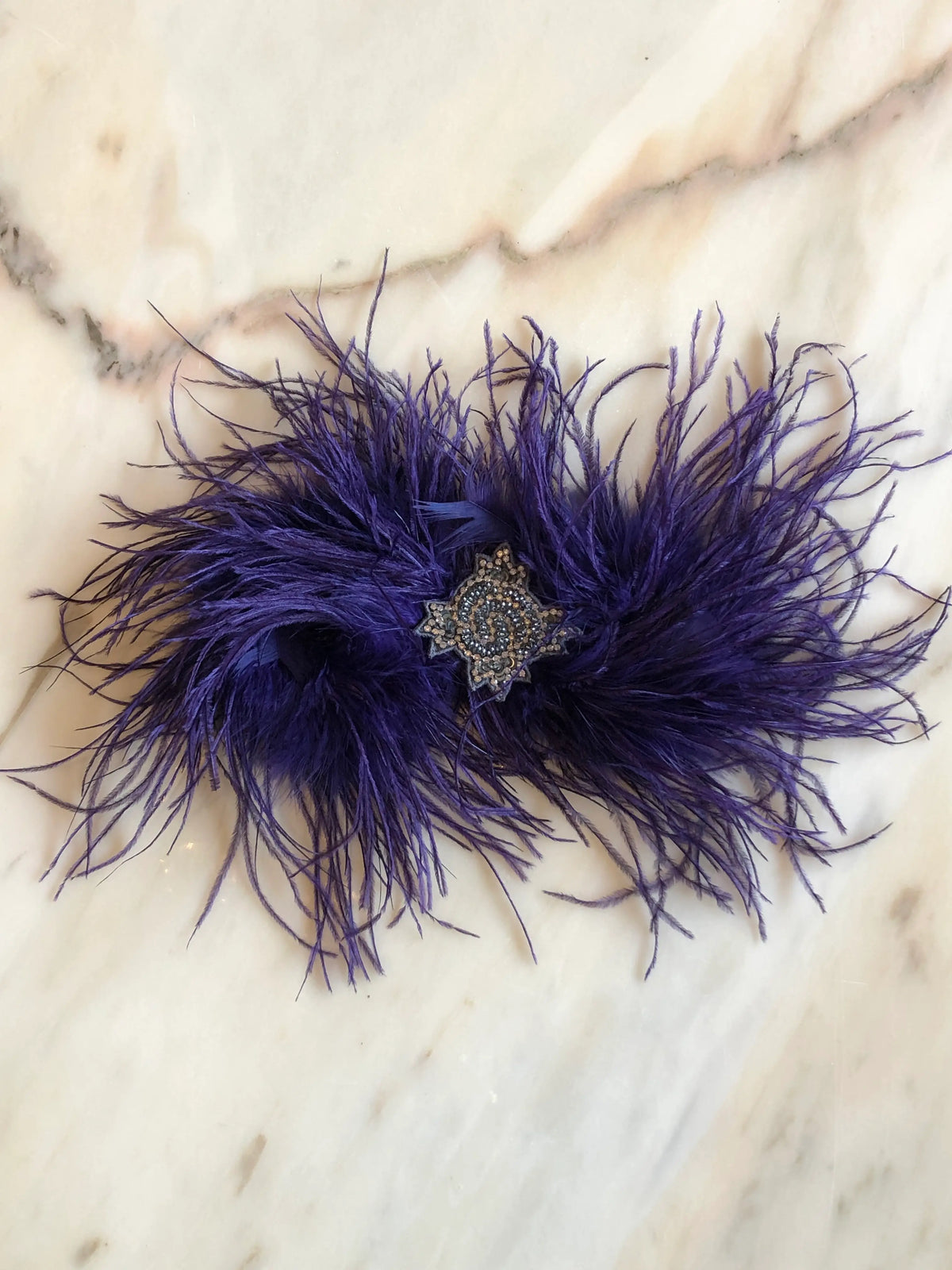 Broochella handmade Purple Feather Bow Brooch featuring dramatic ostrich feathers and an ornate jewelled centre, an oversized designer statement brooch photographed on a white marble background.