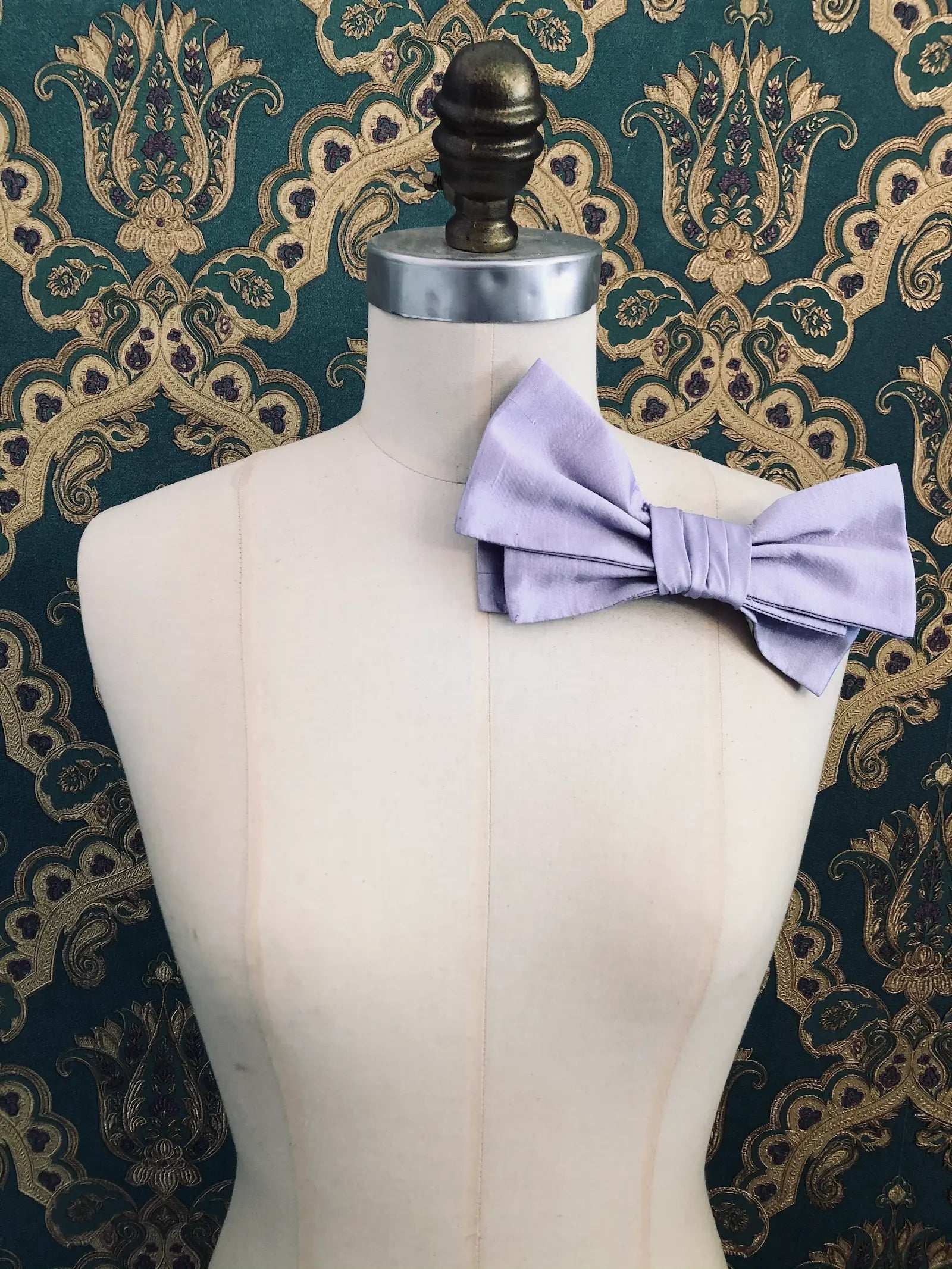 Broochella Marie Antoinette handmade, designer silk bow brooch in purple. It is pinned to a mannequin. The background is green and gold damask wallpaper.
