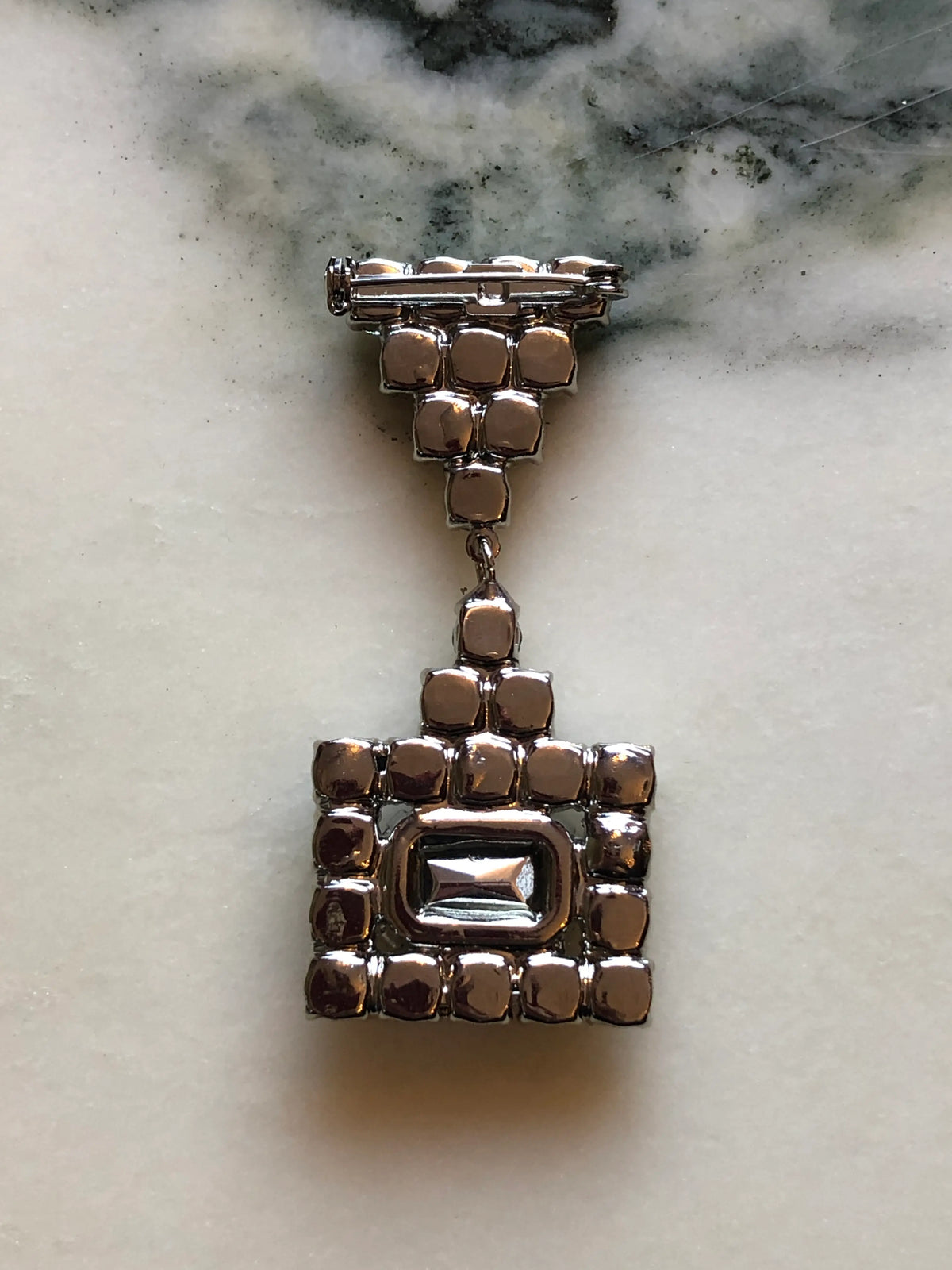 The back view of Broochella Raffaella Vintage Red Crystal Drop Brooch featuring a rectangular ruby-red faceted stone framed by clear crystals with an articulated crystal drop, photographed on a cream and grey marble background.