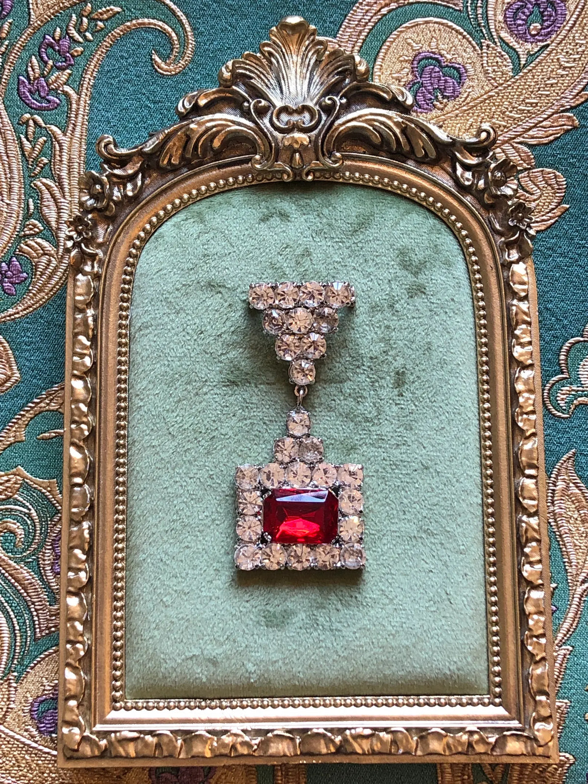Broochella Raffaella Vintage Red Crystal Drop Brooch featuring a rectangular ruby-red faceted stone framed by clear crystals with an articulated crystal drop. It is photographed on a green velvet tray with an ornate gold frame. The background is green and gold damask wallpaper.
