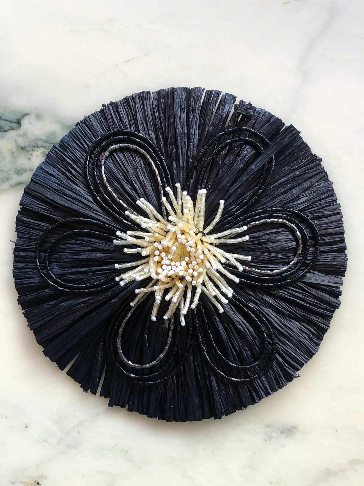 Broochella Men's Raffia Bloom Brooch Pin in black with sculptural raffia fringe, black beaded flower petals and an ivory beaded centre, photographed on a white marble background.