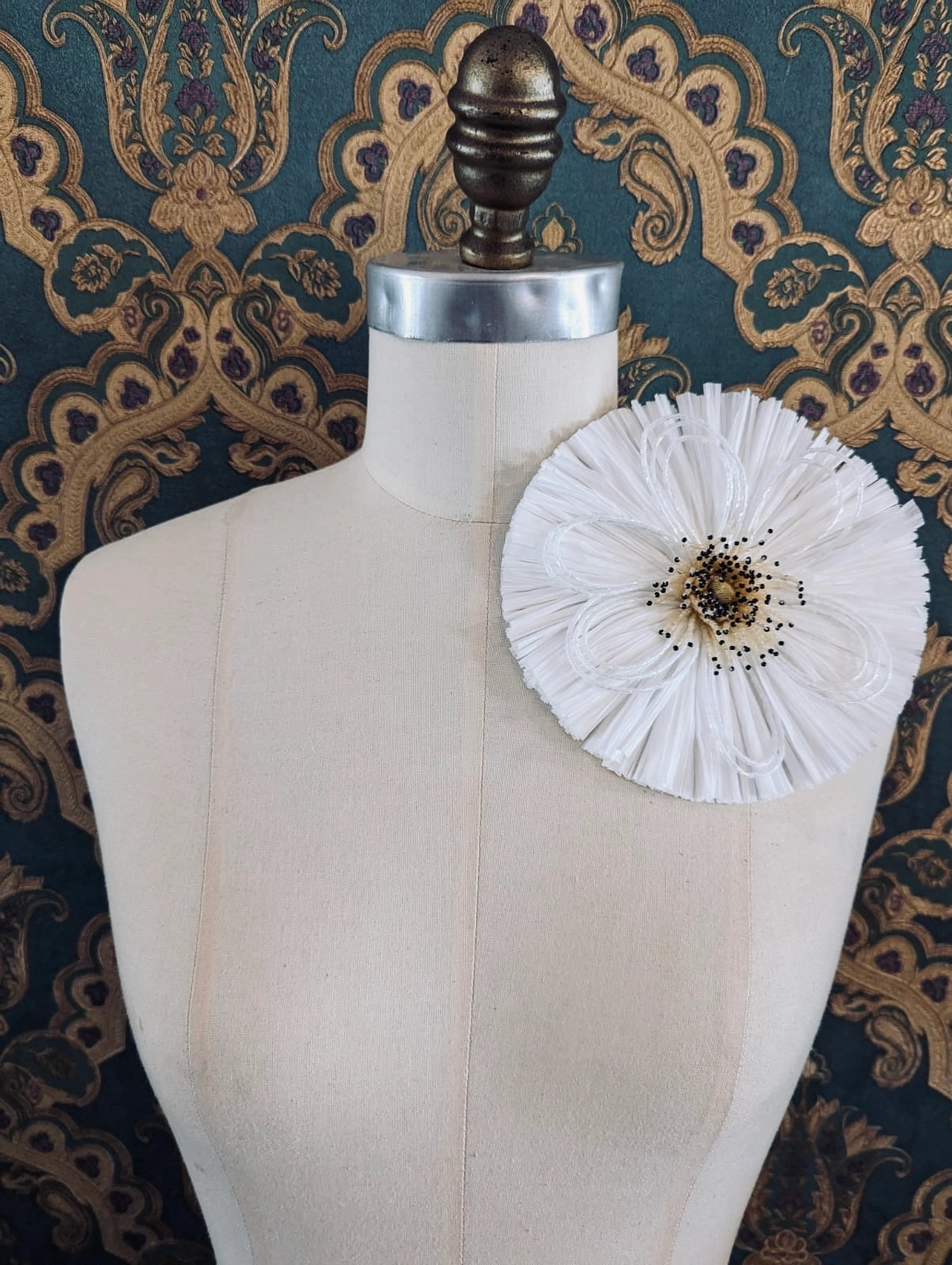 Broochella Raffia Bloom white  handmade raffia flower brooch with hand beaded centre. Pinned to a mannequin. The background is green and gold damask wallpaper