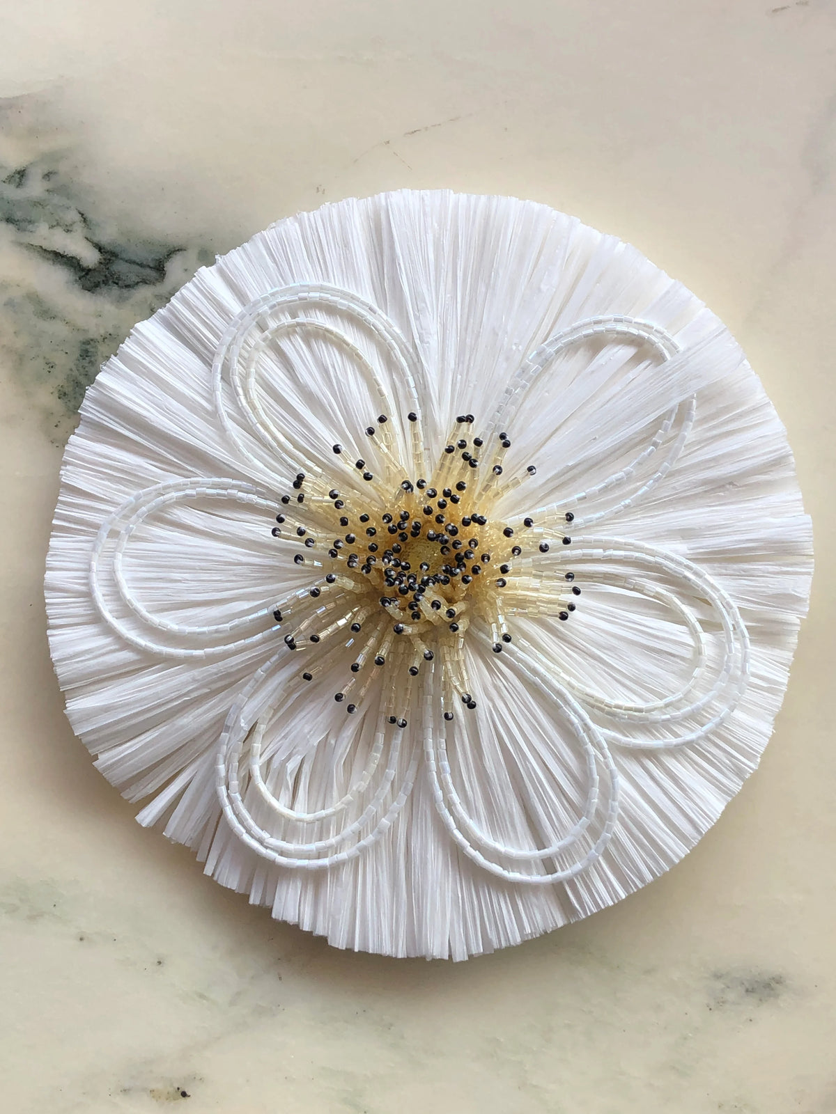 Broochella Raffia Bridal Flower Brooch in white, featuring a sculptural raffia bloom with looping white beadwork and a beaded statement centre, photographed on a pale cream marble background.