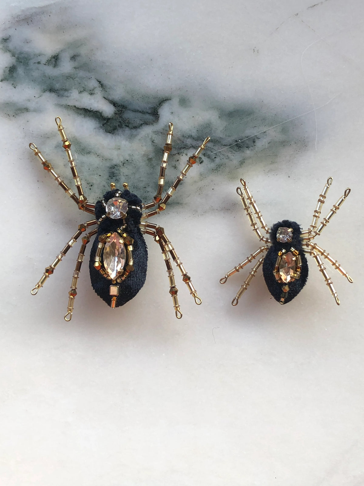 Broochella Ragno e Ragnetto Men's Spider Brooch Pin Set featuring two black textile spiders embellished with gold-tone beadwork and faceted crystal details. Pictured on a white marble surface