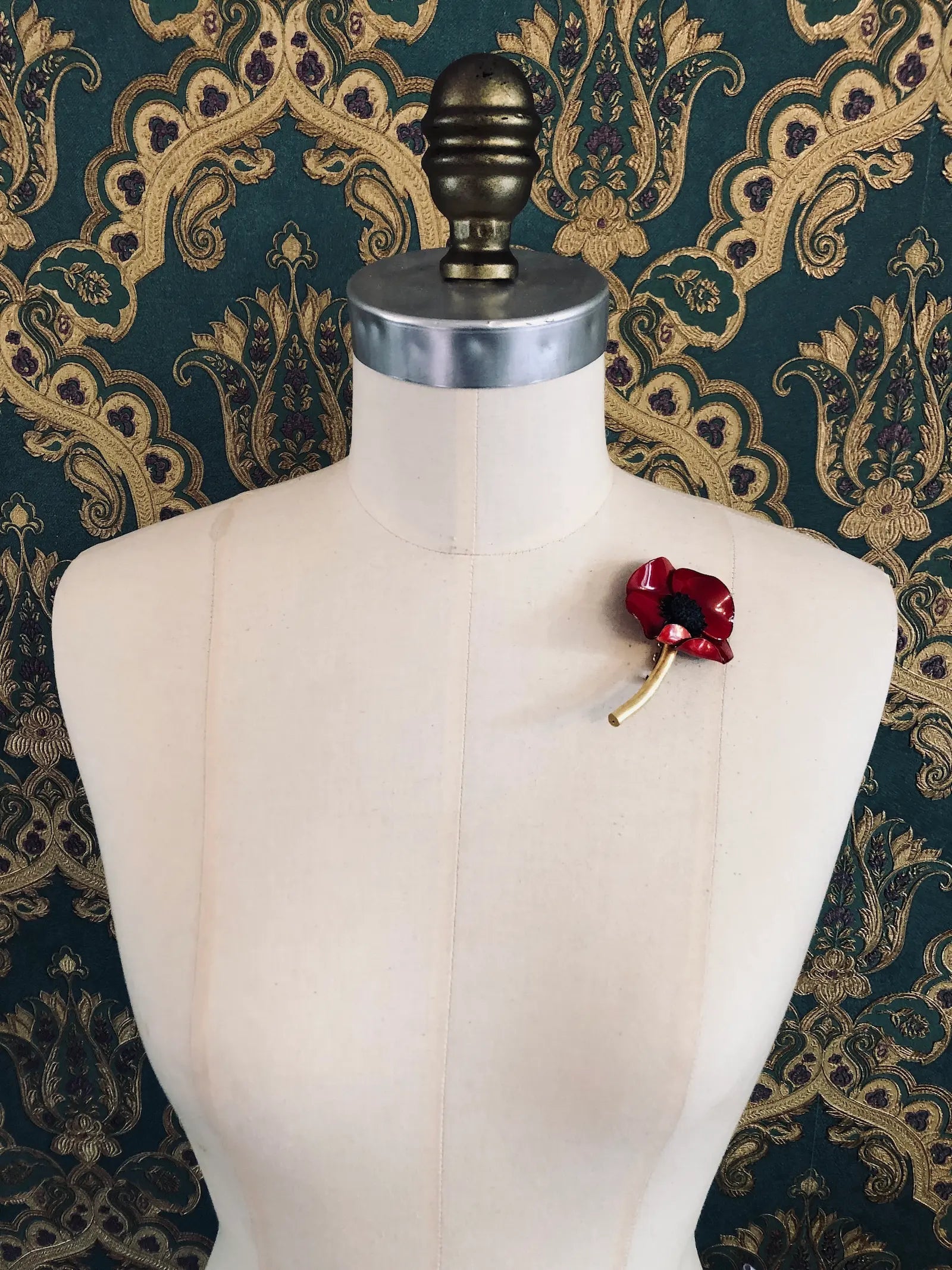 Broochella x Fabrice Paris red moulded acetate poppy flower brooch in red. It is pinned to a mannequin. The background is green and gold damask wallpaper. 