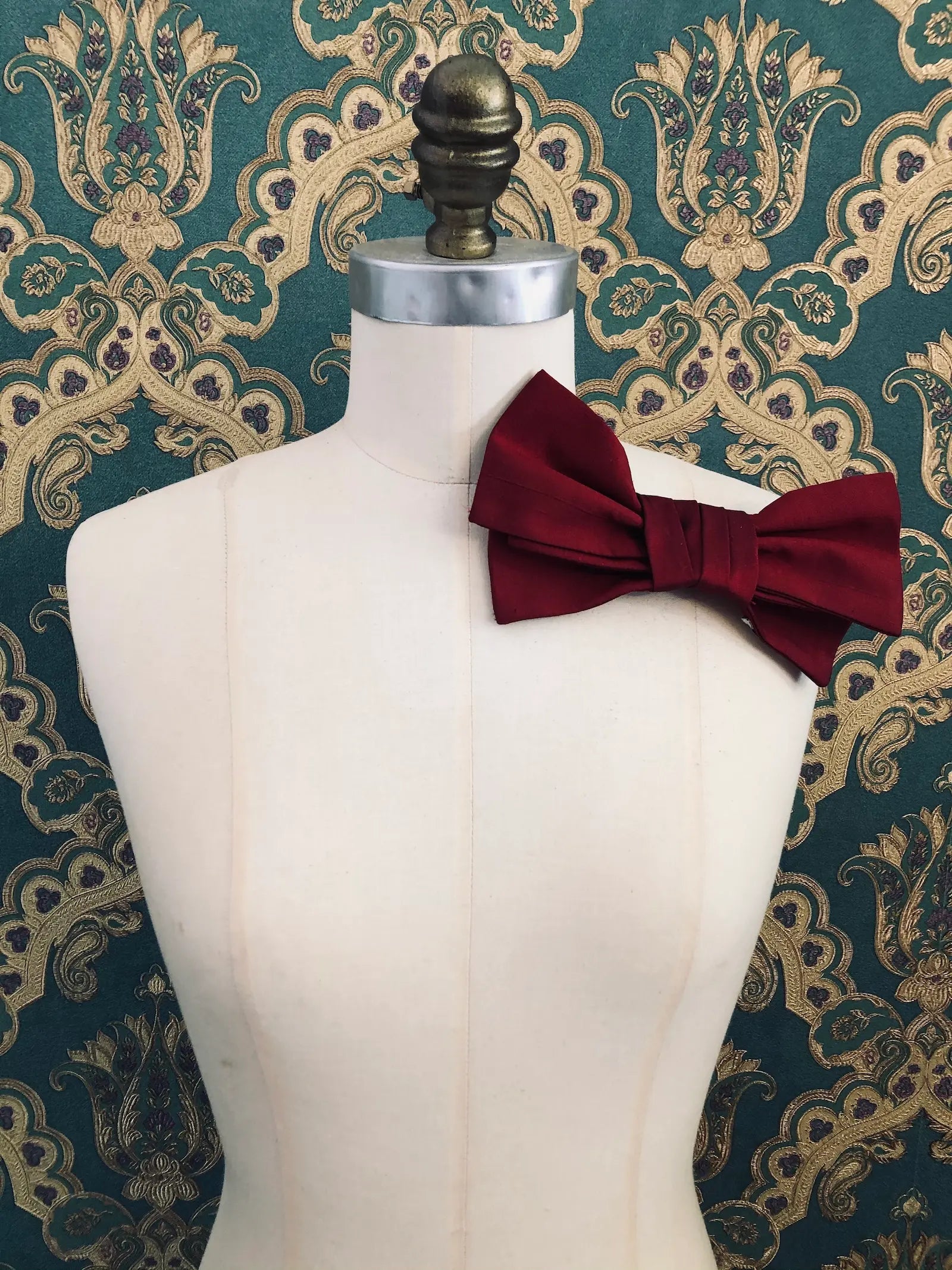 Broochella Marie Antoinette handmade, designer silk bow brooch in red. It is pinned to a mannequin. The background is green and gold damask wallpaper.