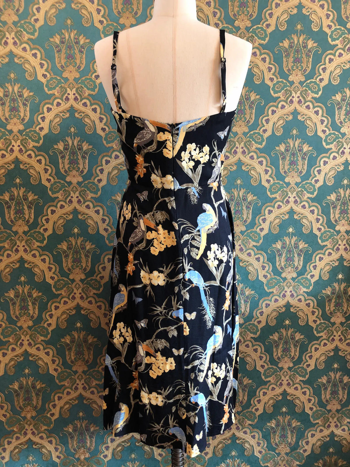 The back view Broochella Reformation black midi dress with colourful tropical birds, butterflies and floral print, size 10. Pictured on a mannequin. The background is green and gold damask wallpaper