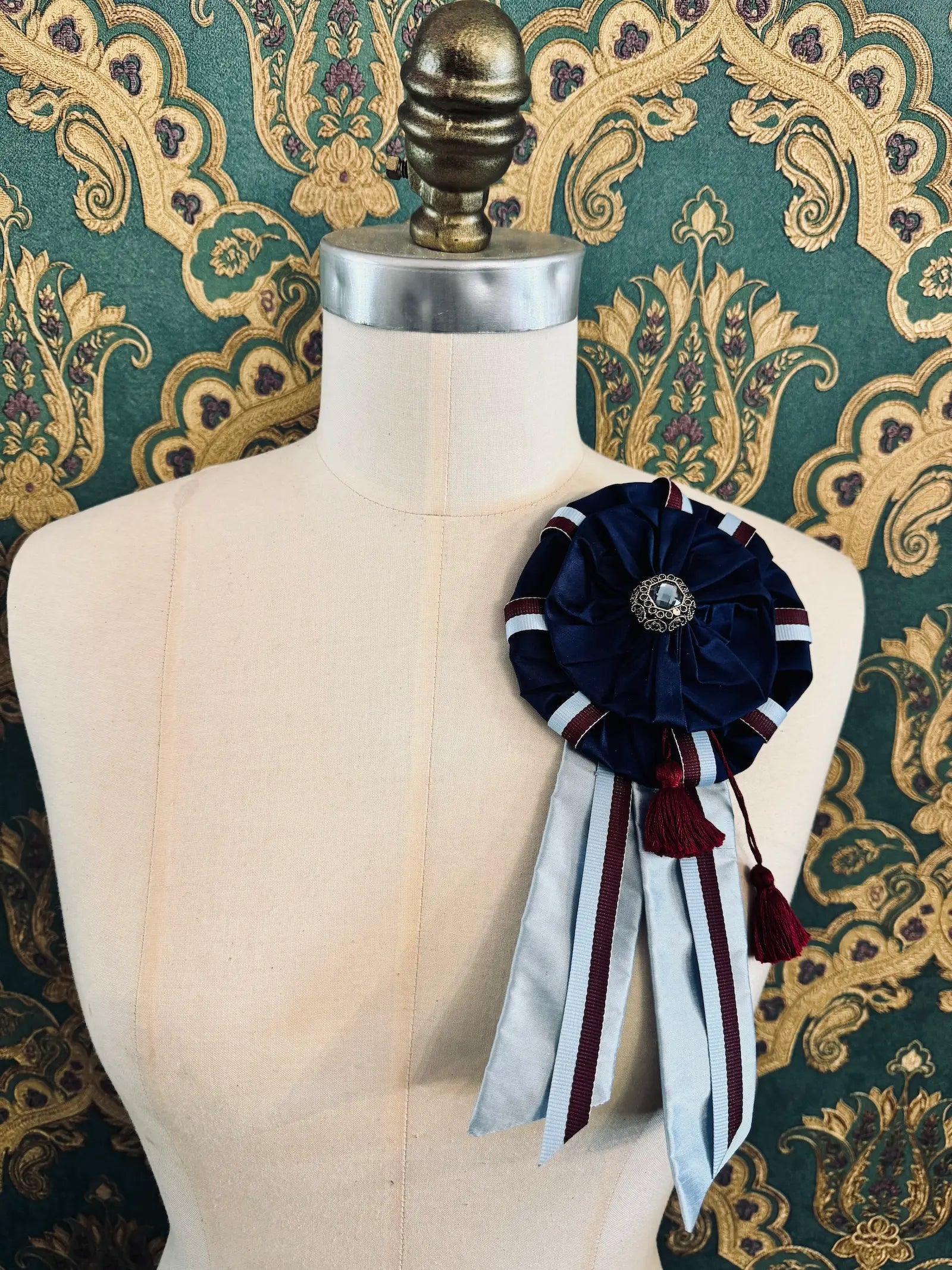 Broochella regalia silk ribbon brooch with vintage ribbon trim in navy. Pictured on a mannequin. The background is green and gold damask wallpaper.