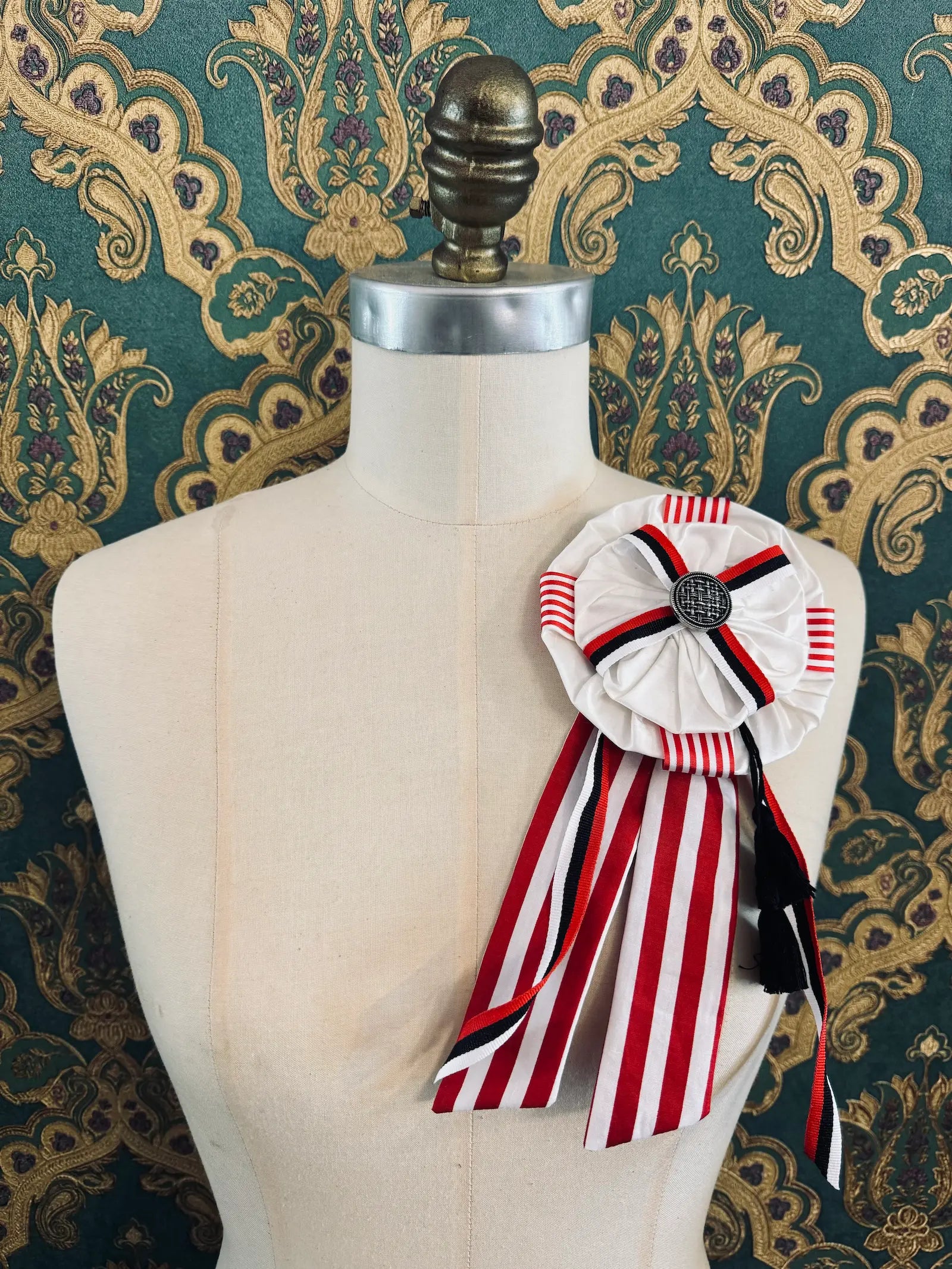 Broochella regalia silk ribbon brooch with vintage ribbon trim in red, white and black. Pictured on a mannequin. The background is green and gold damask wallpaper.