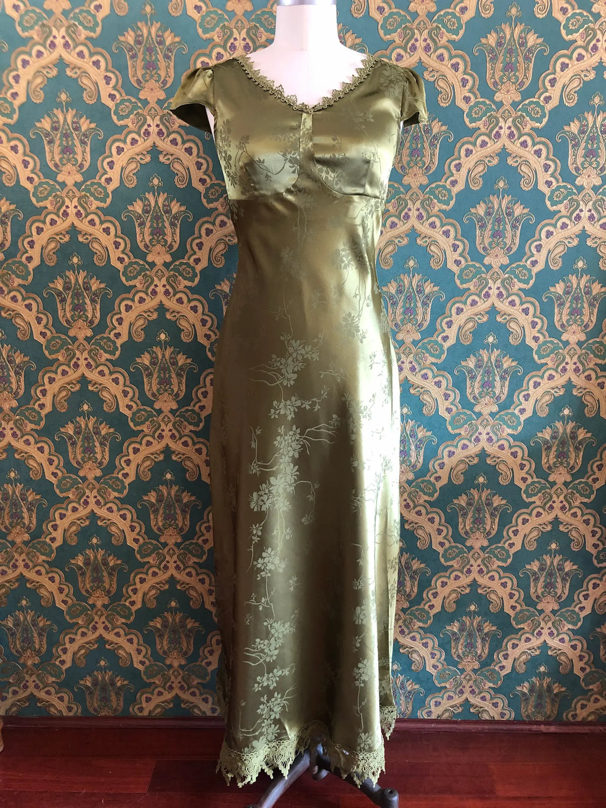 Broochella Rihoas olive-green floral jacquard satin maxi dress with lace trim and an open tie back, size Medium. Pictured on a mannequin. The background is green and gold damask wallpaper