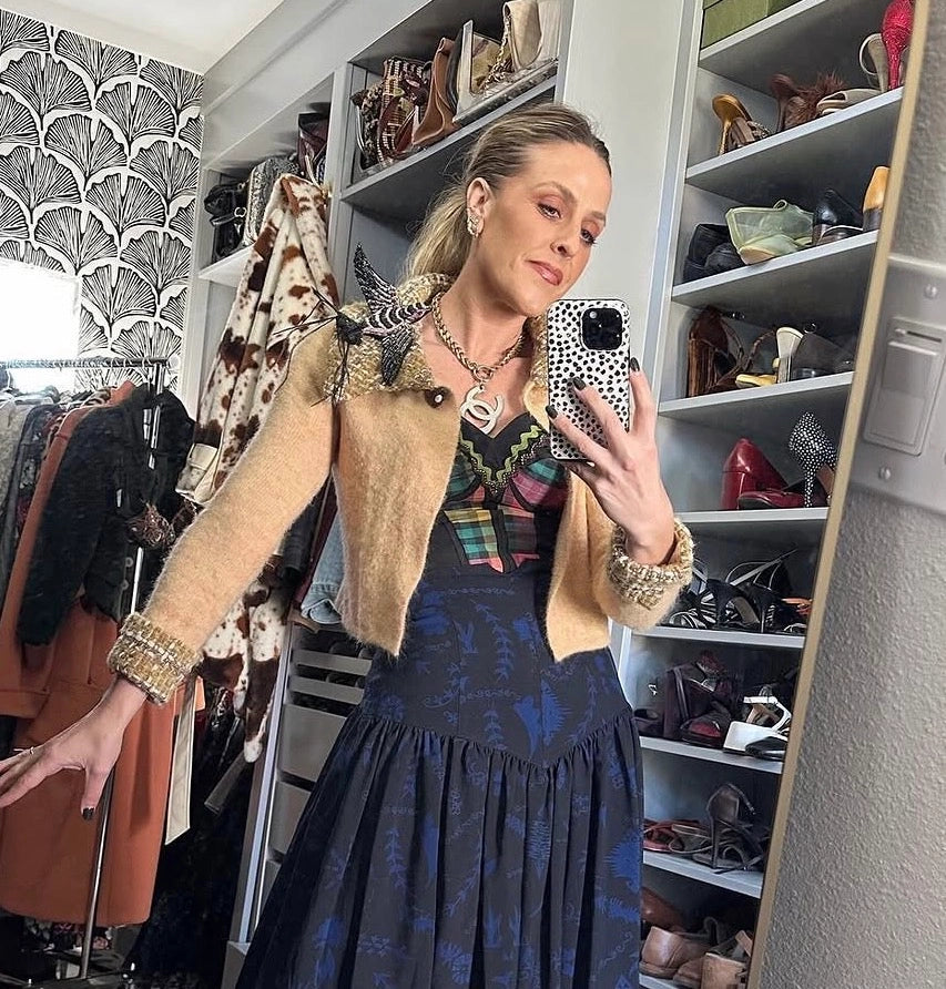 A woman takes a mirror selfie in a walk-in closet, wearing a beige cropped jacket with leopard-print trim over a dark blue patterned dress. Shelves in the background are filled with shoes and other clothing. She is wearing the Broochella Rondine hand beaded bird brooch in blak and pink. 